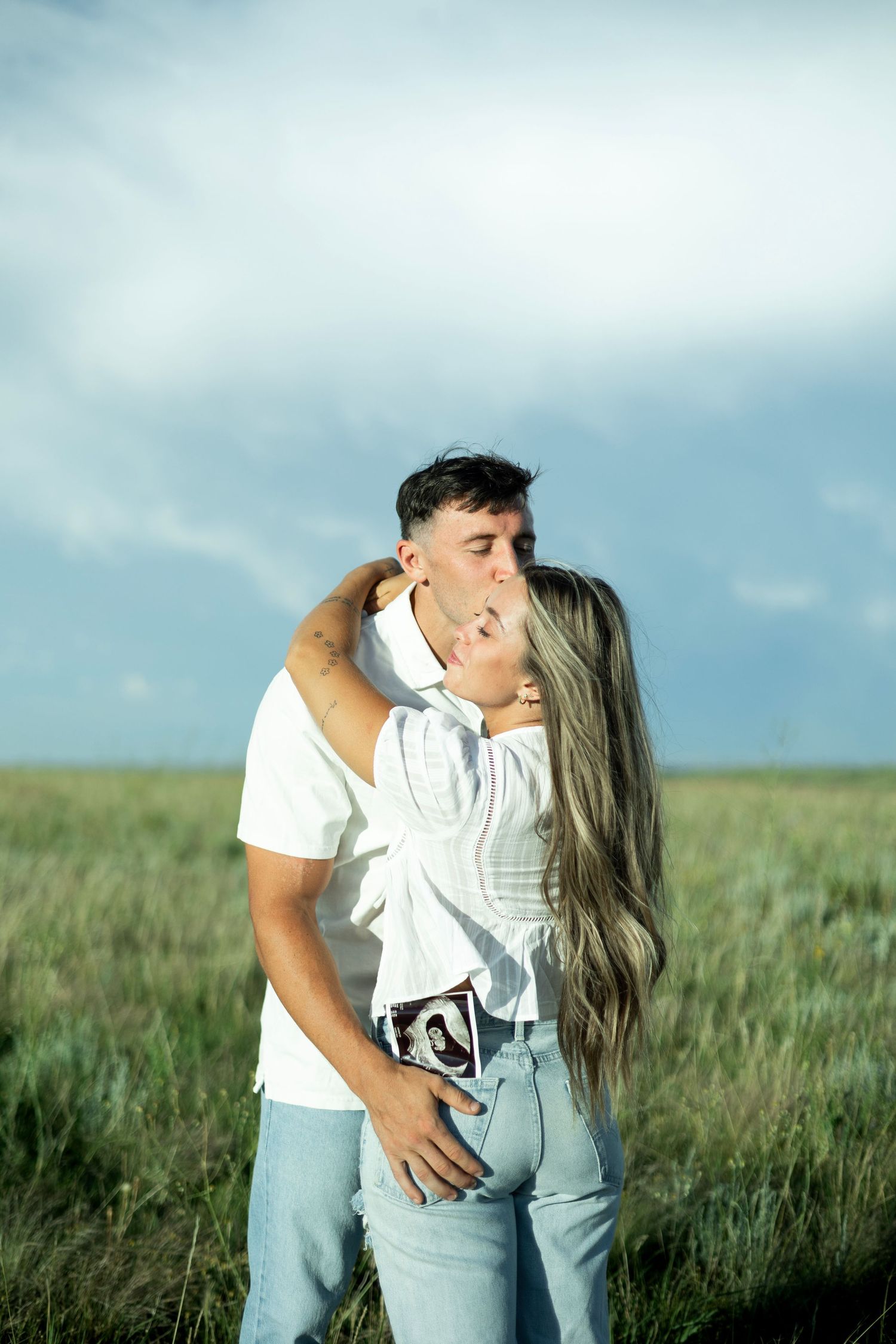 A tender kiss shared between two people in casual clothing amidst a grassy field under a cloudy sky.