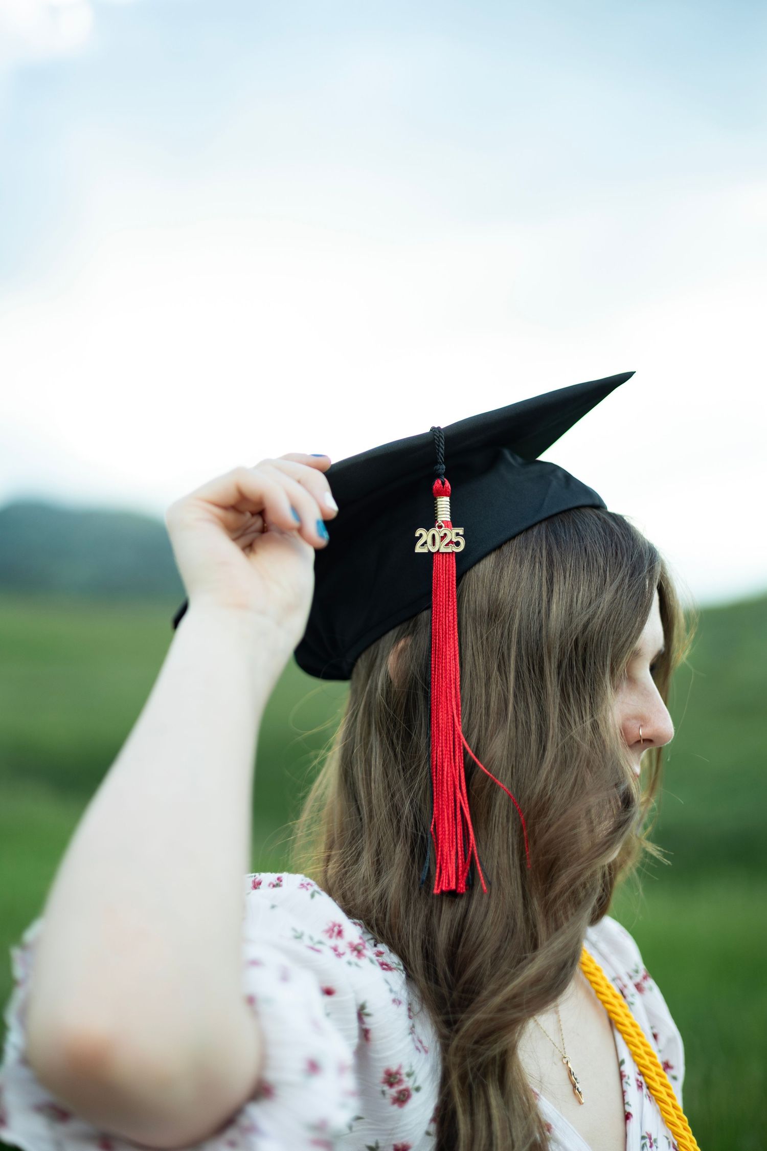 Graduate wearing a mortarboard cap with red tassel and floral dress poses in an outdoor field with rolling hills.
