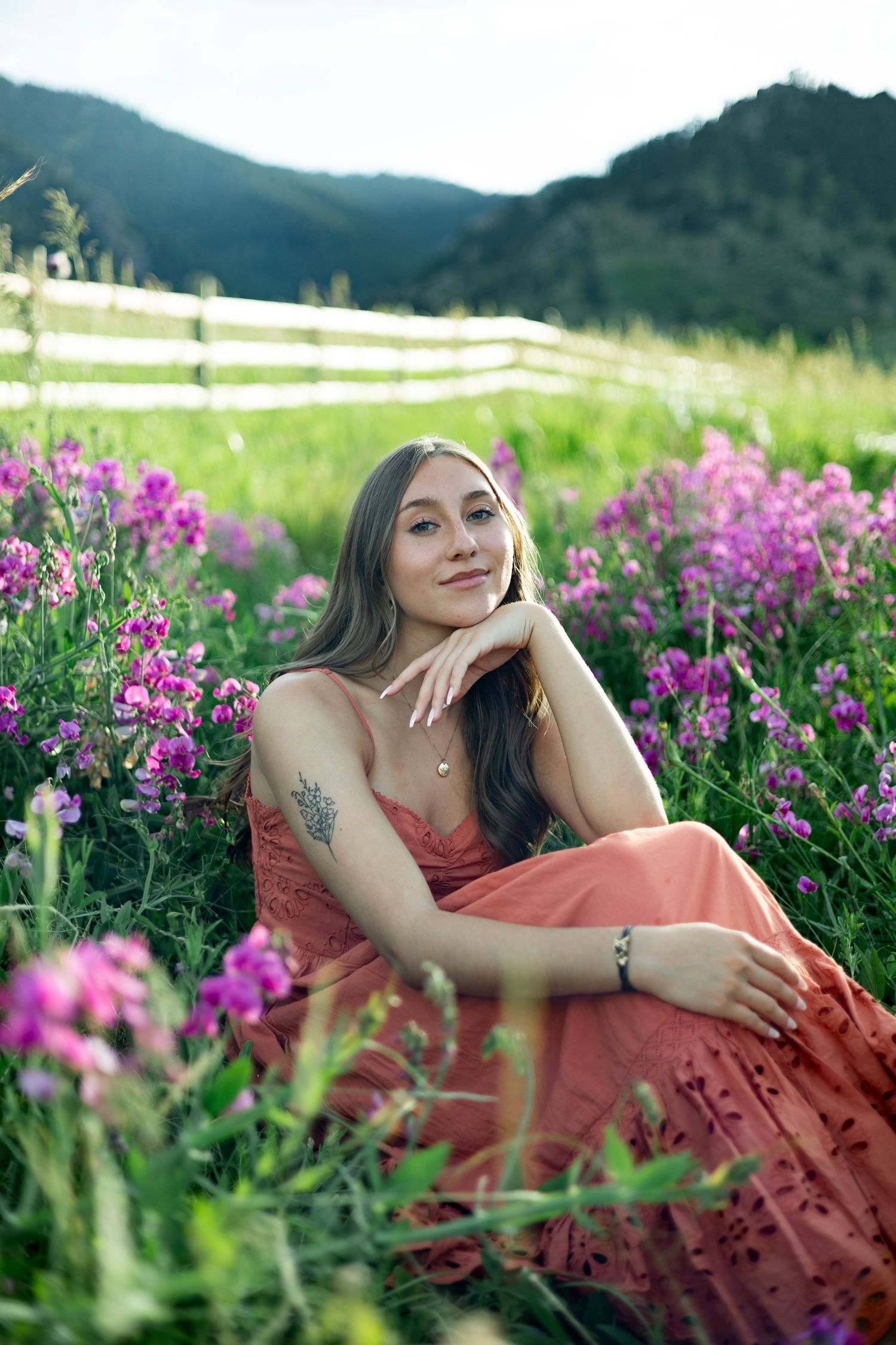 A person in a coral dress poses gracefully among vibrant purple wildflowers in a mountain meadow setting.