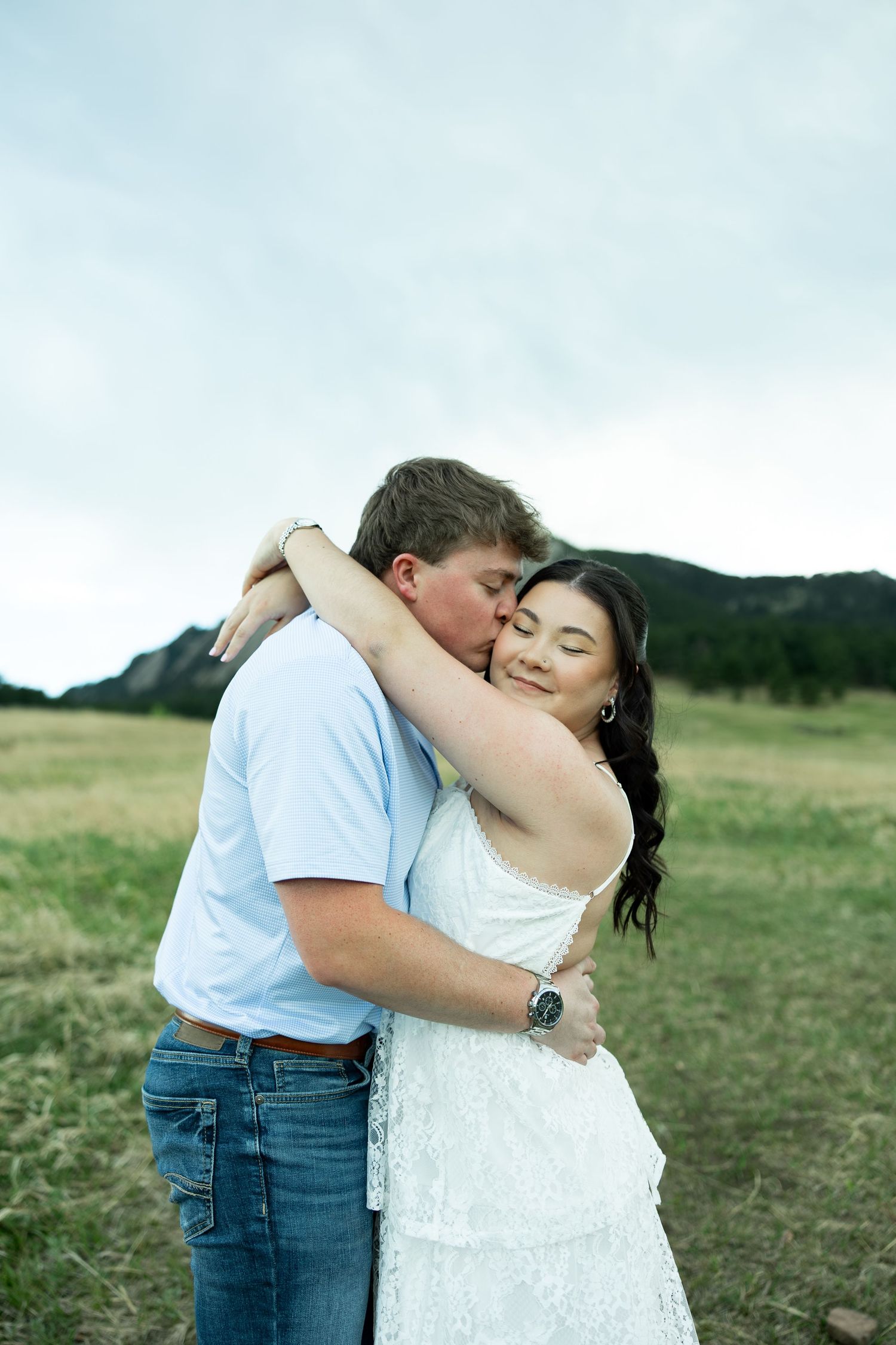 Couple embracing in a green meadow with mountain backdrop, woman in white dress smiling as man kisses her cheek.