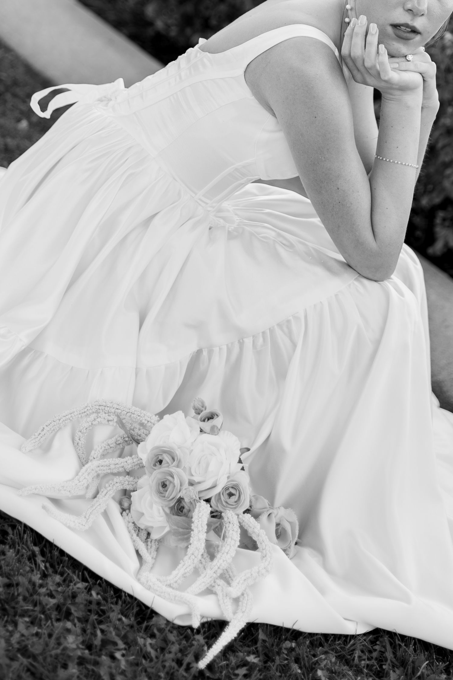 Black and white photo of a bride in a white dress sitting on grass with a floral bouquet beside her.