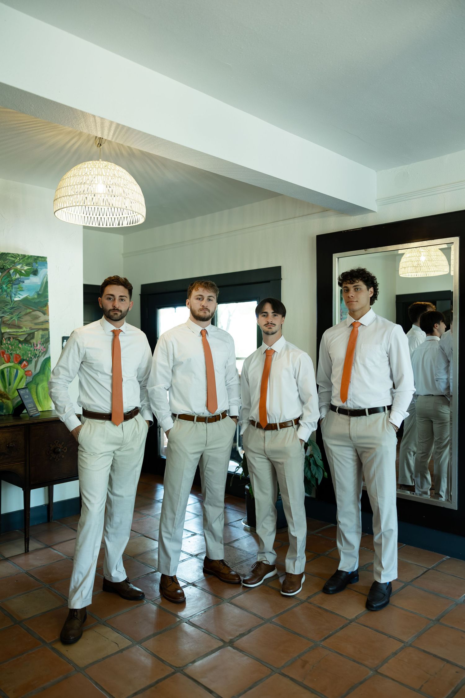 Four groomsmen in white shirts and orange ties pose together in an elegant room before a wedding ceremony.