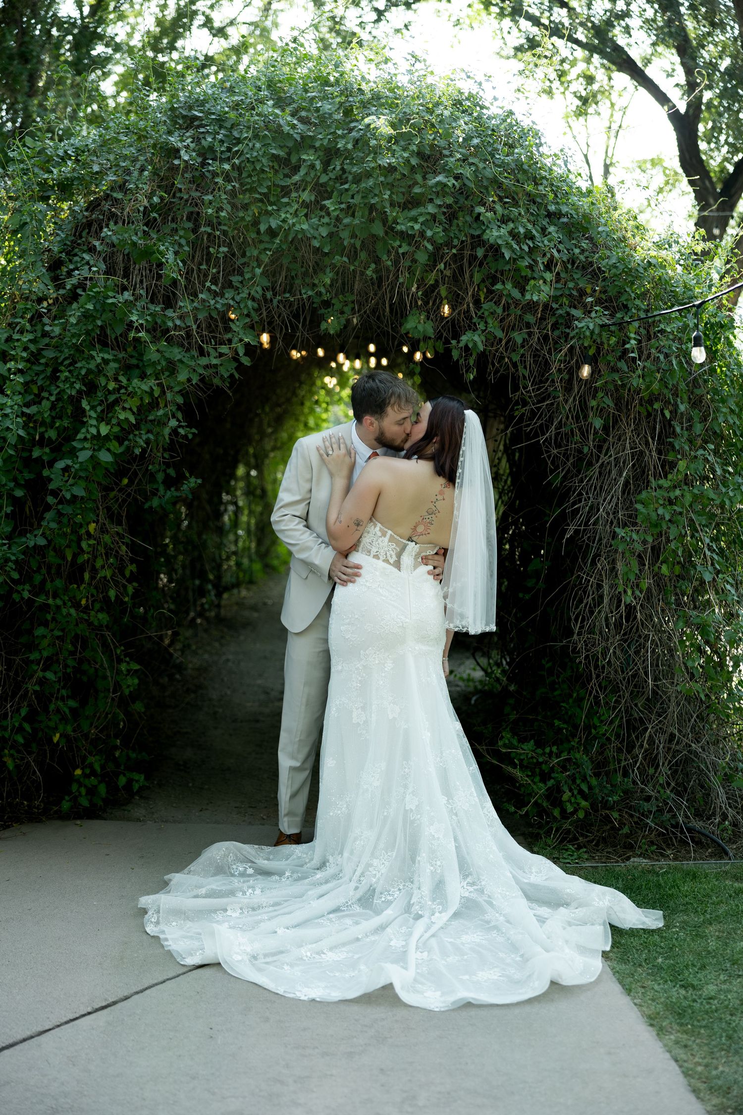 Bride and groom share a romantic kiss under a lush ivy archway with string lights at their wedding.