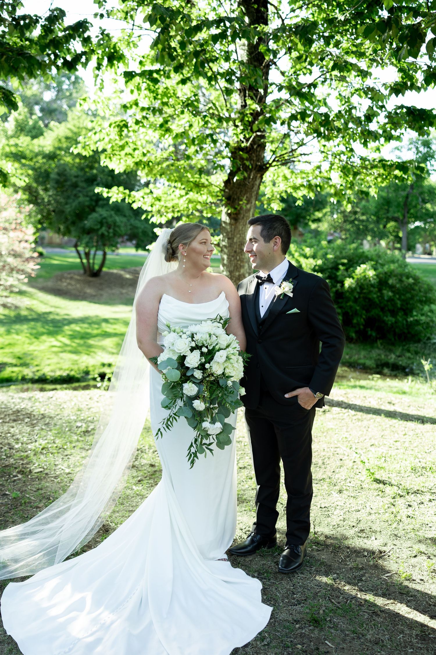 Bride in white gown with veil and groom in black tuxedo share a romantic moment outdoors under green trees.