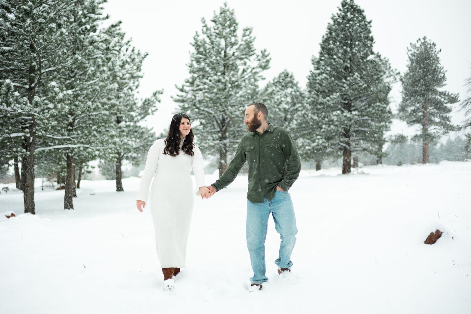 A couple walks hand in hand through a snow-covered pine forest.