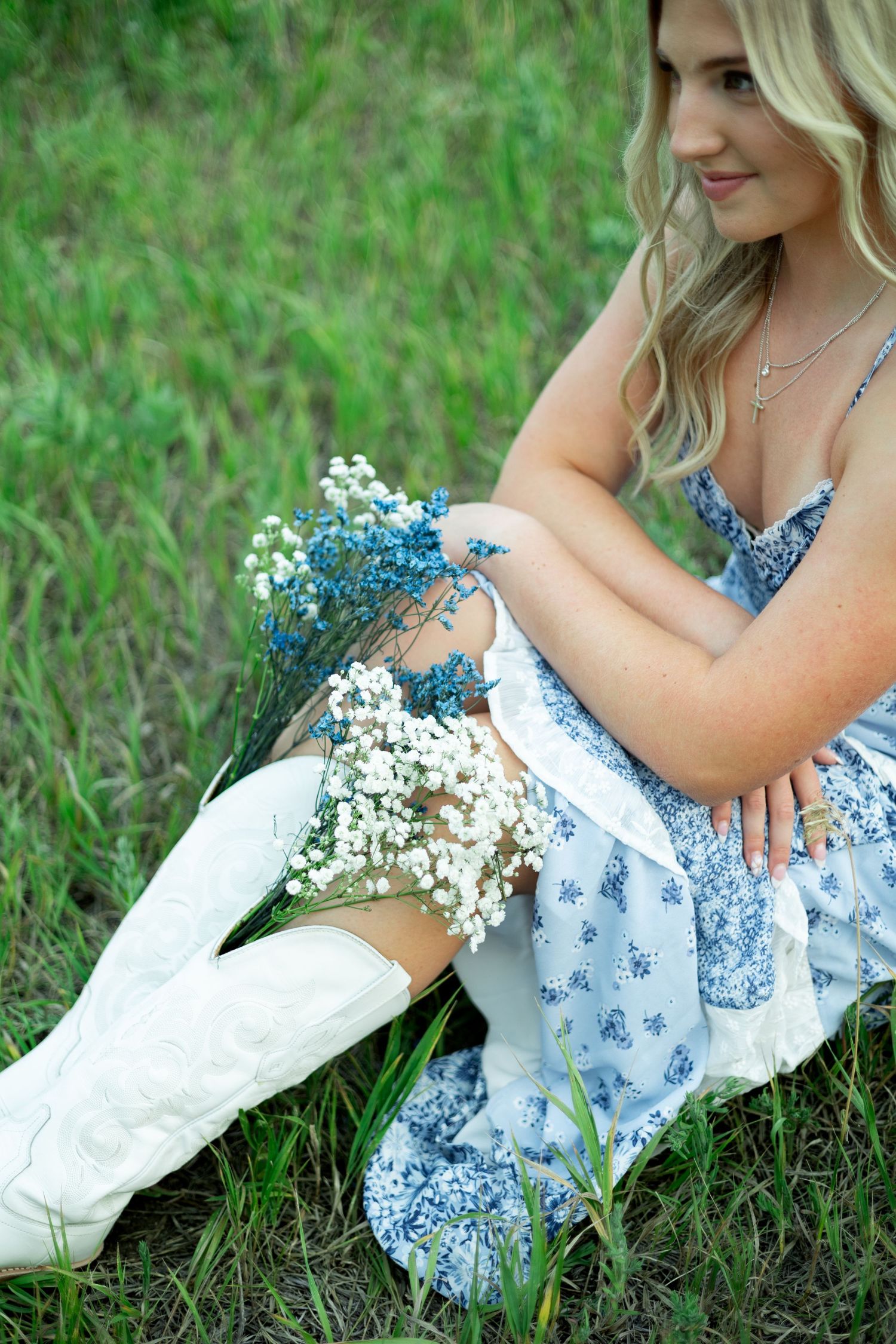 Person wearing a blue floral dress sits in grass holding white cowboy boots and blue wildflowers.