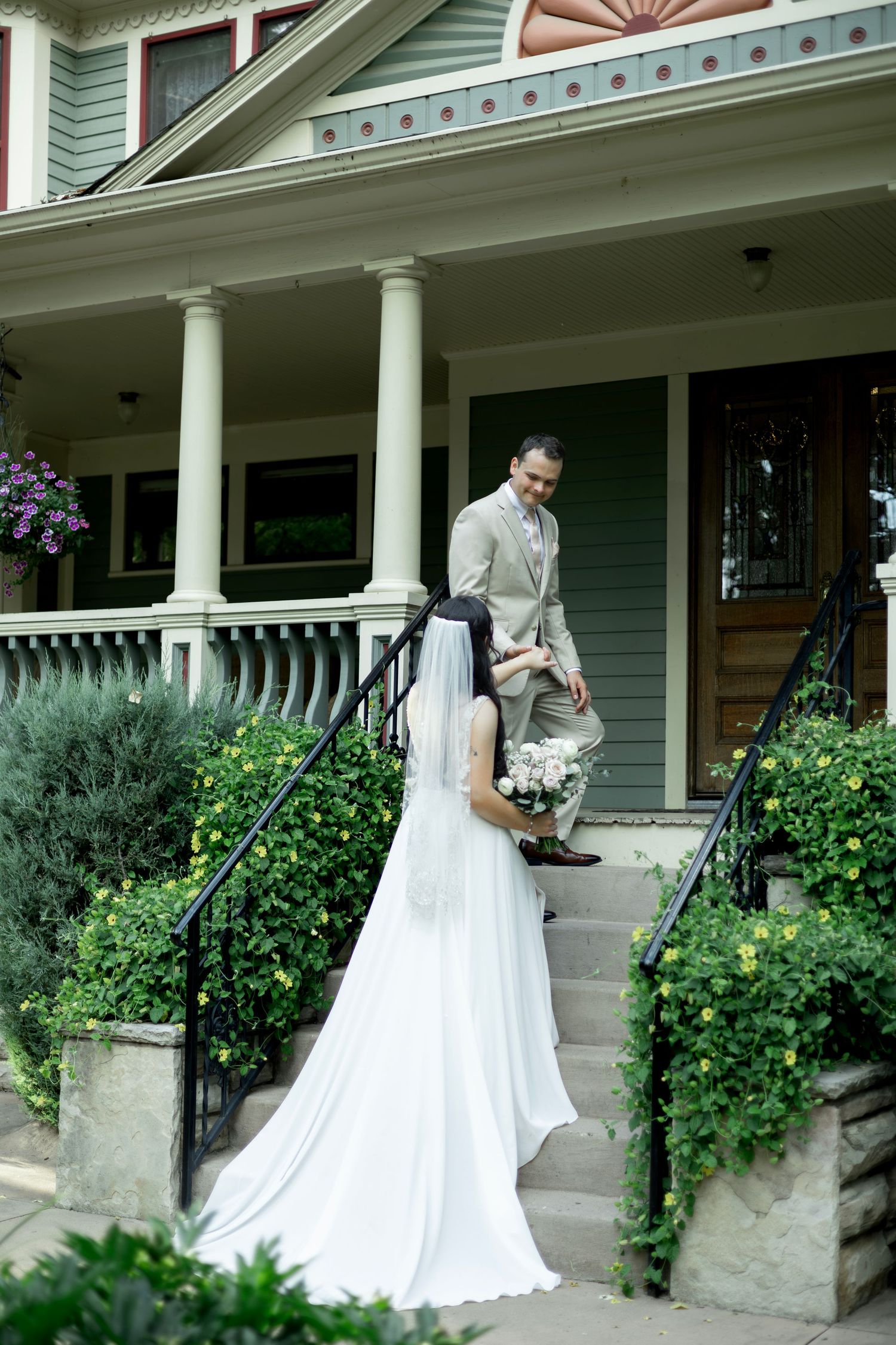 Bride in white gown with veil approaches groom in tan suit on steps of elegant Victorian home.