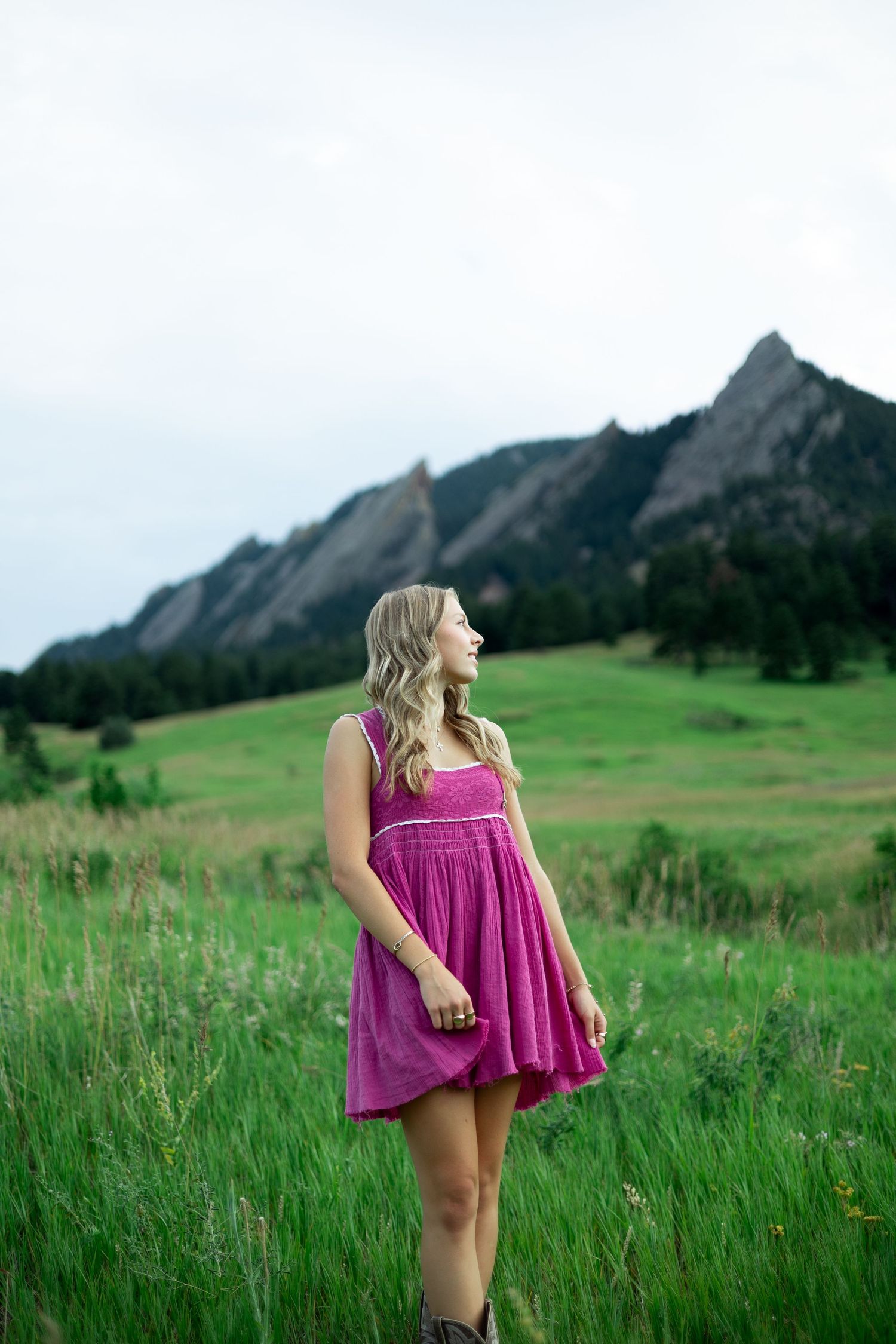 Person in a flowy magenta dress poses against a scenic backdrop of rolling green hills and dramatic mountain peaks.