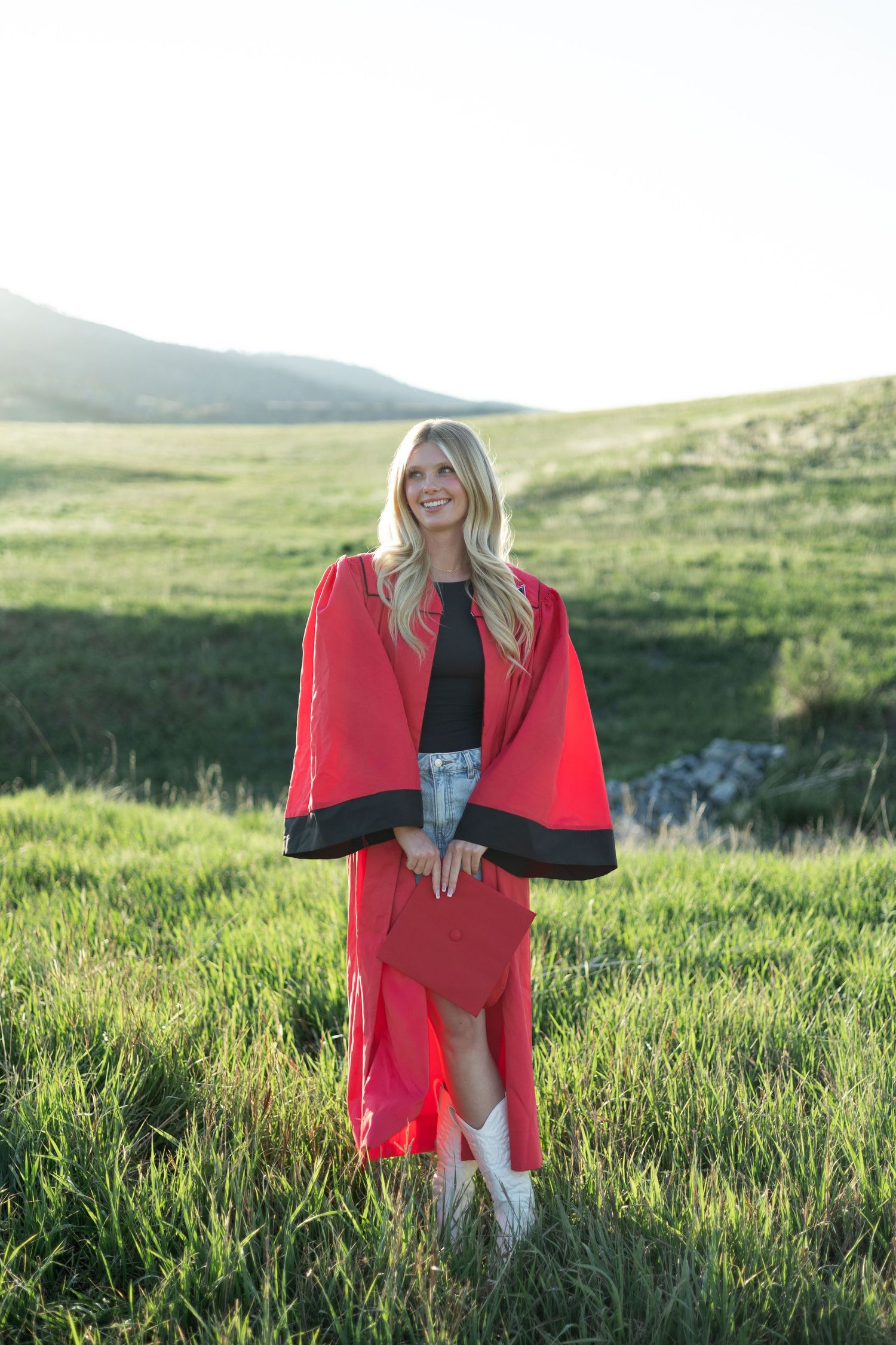 Stylish red and black cape outfit worn in sunny green field against mountain backdrop at golden hour.