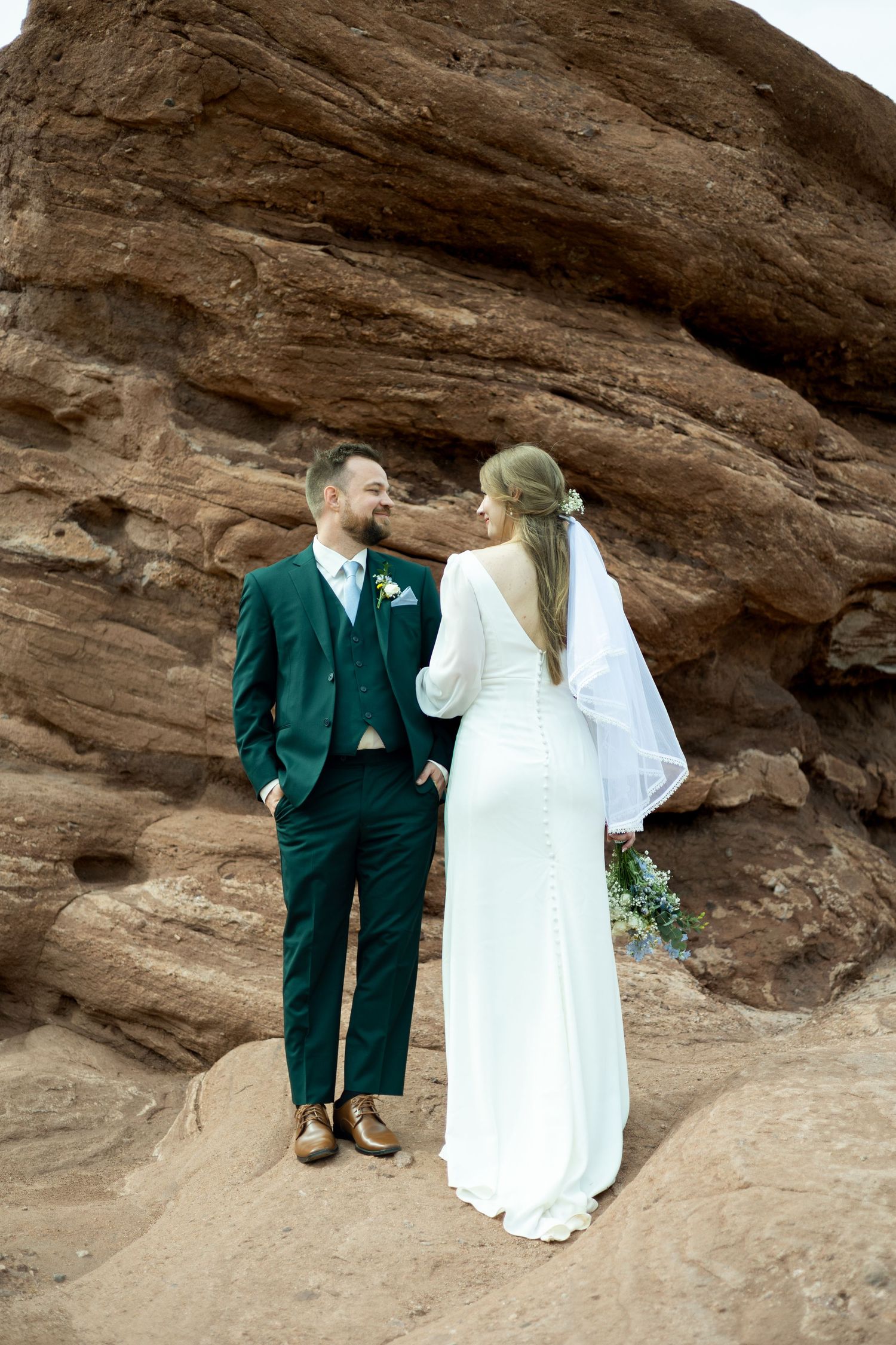 A couple in wedding attire stands against dramatic red rock formations in an outdoor desert setting.