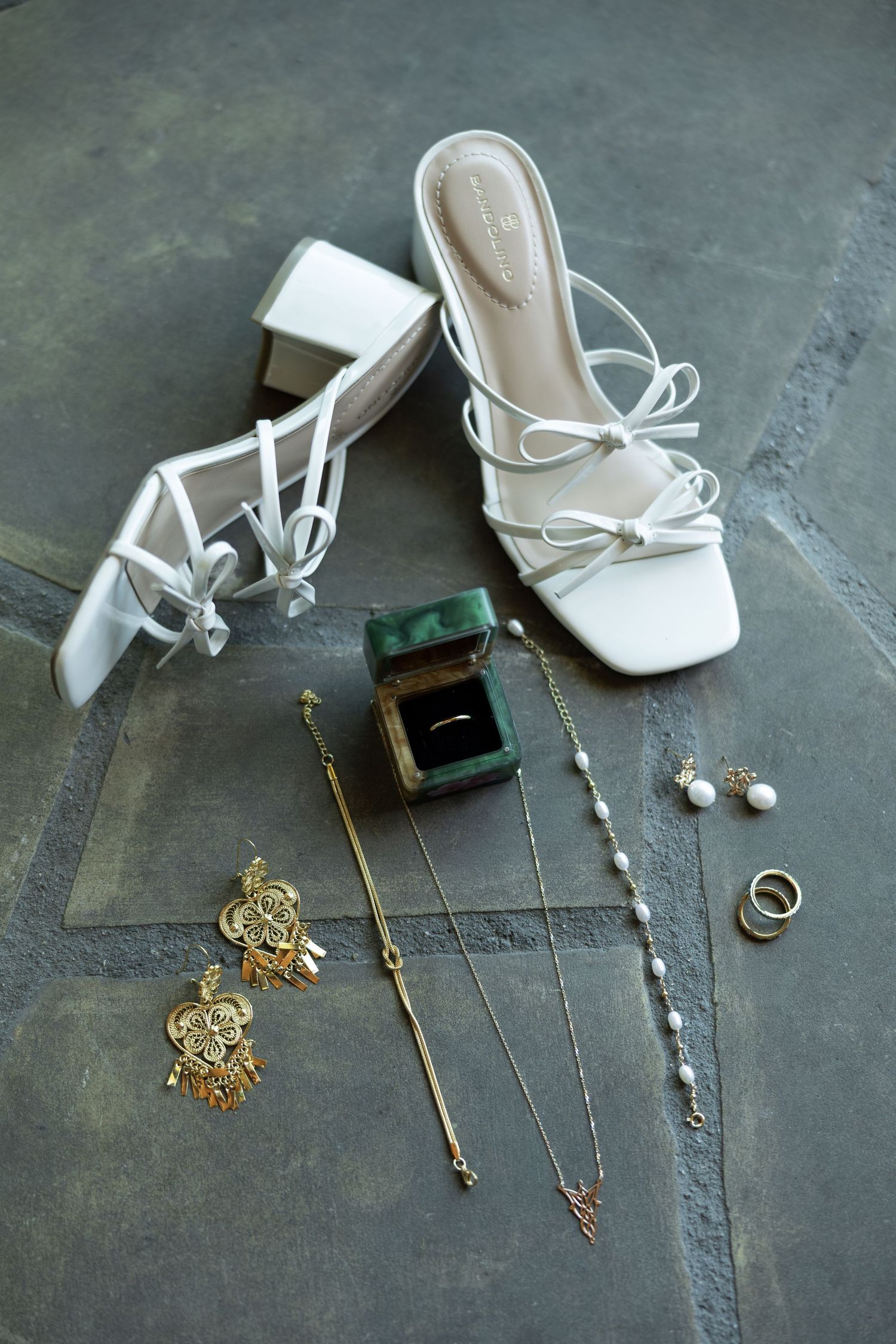 White lace-up block heel sandals with gold jewelry, pearl necklace, earrings, and a green ring box on gray stone.