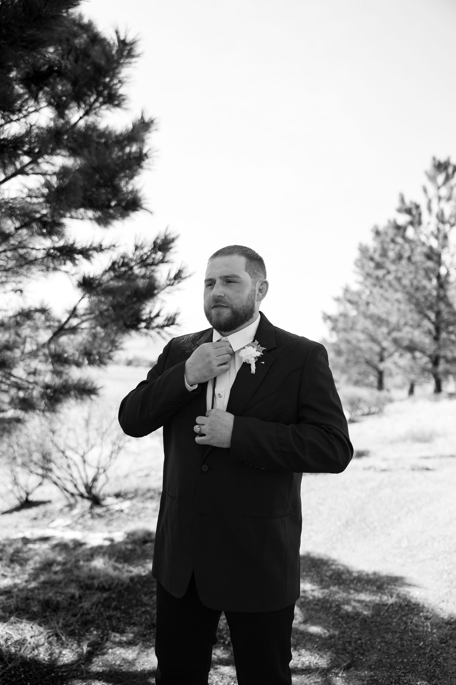 Groom in black suit adjusting his tie outdoors among trees in a black and white wedding portrait.