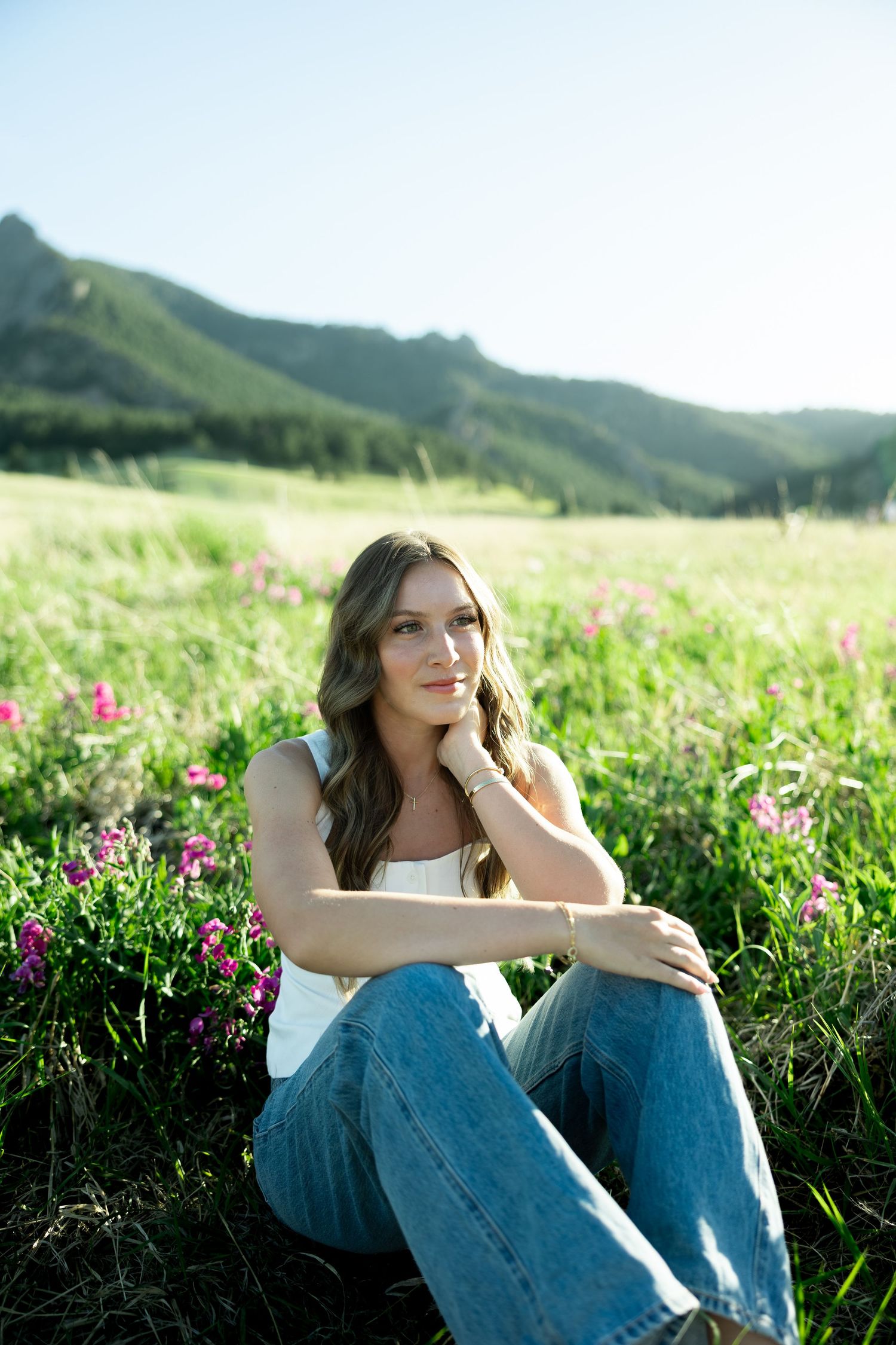 Young woman sitting in a wildflower meadow with mountain views, wearing jeans and white tank top.