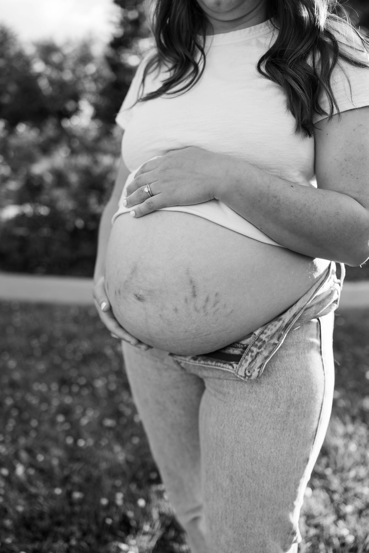 Black and white maternity photo of pregnant woman cradling bare baby bump outdoors, showing stretch marks.