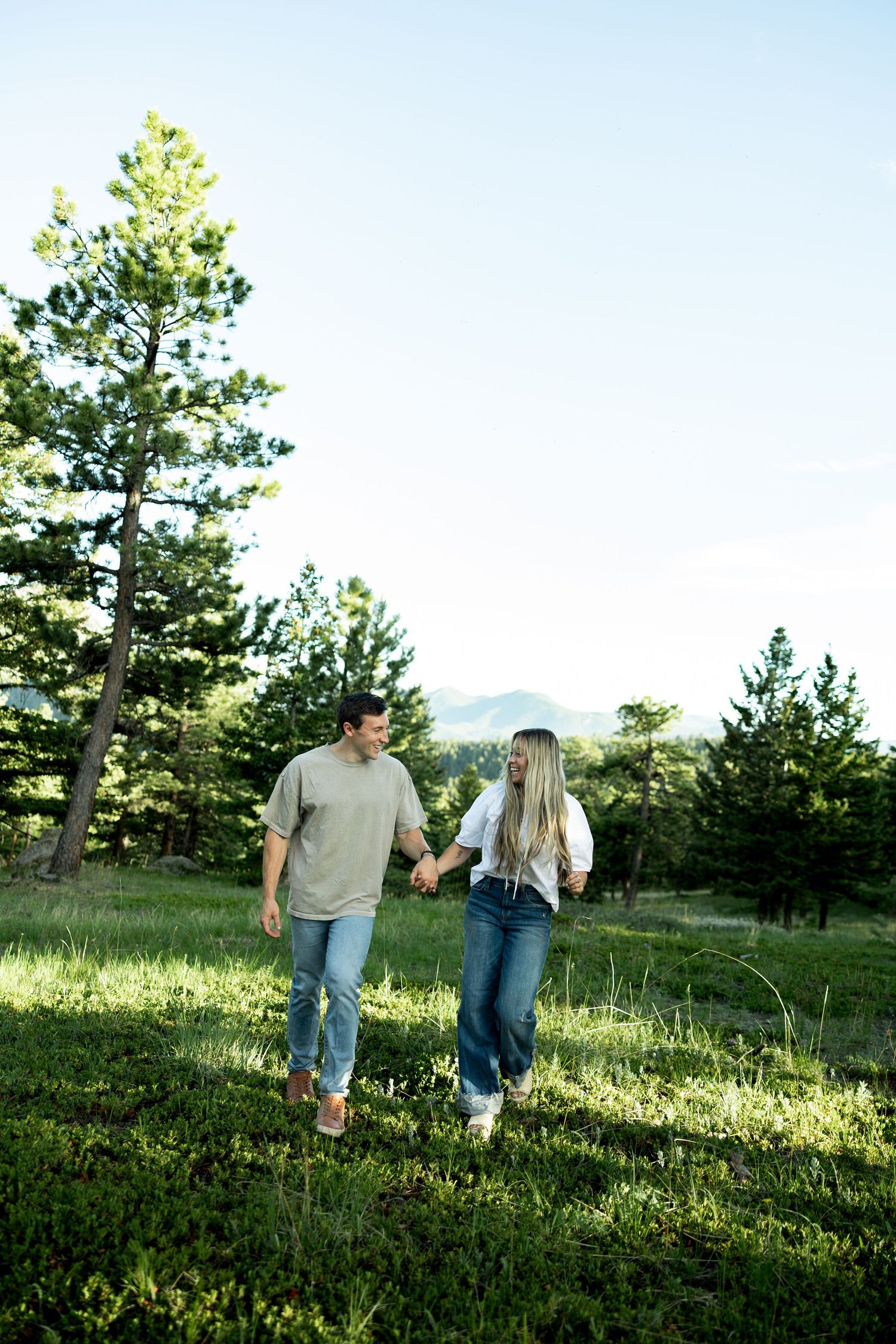 A couple holds hands while walking through a lush green meadow surrounded by tall pine trees and mountain views.