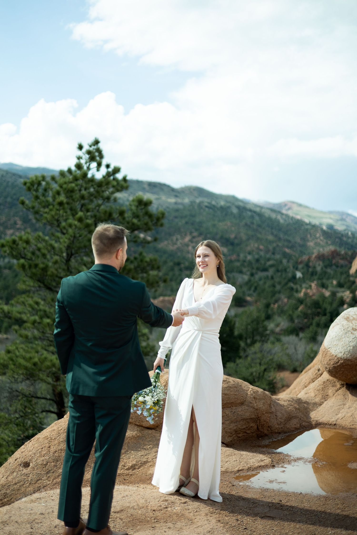 A couple in wedding attire stands on rocky terrain against a scenic mountain backdrop during their outdoor ceremony.