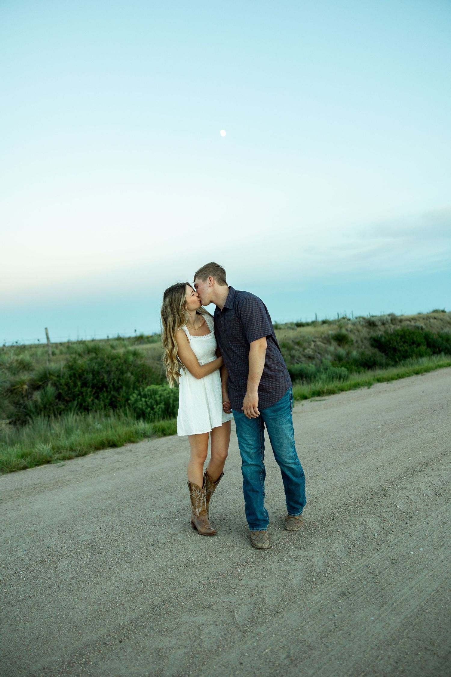 Couple sharing a kiss on a dirt road at dusk, woman in white dress and cowboy boots, man in jeans.