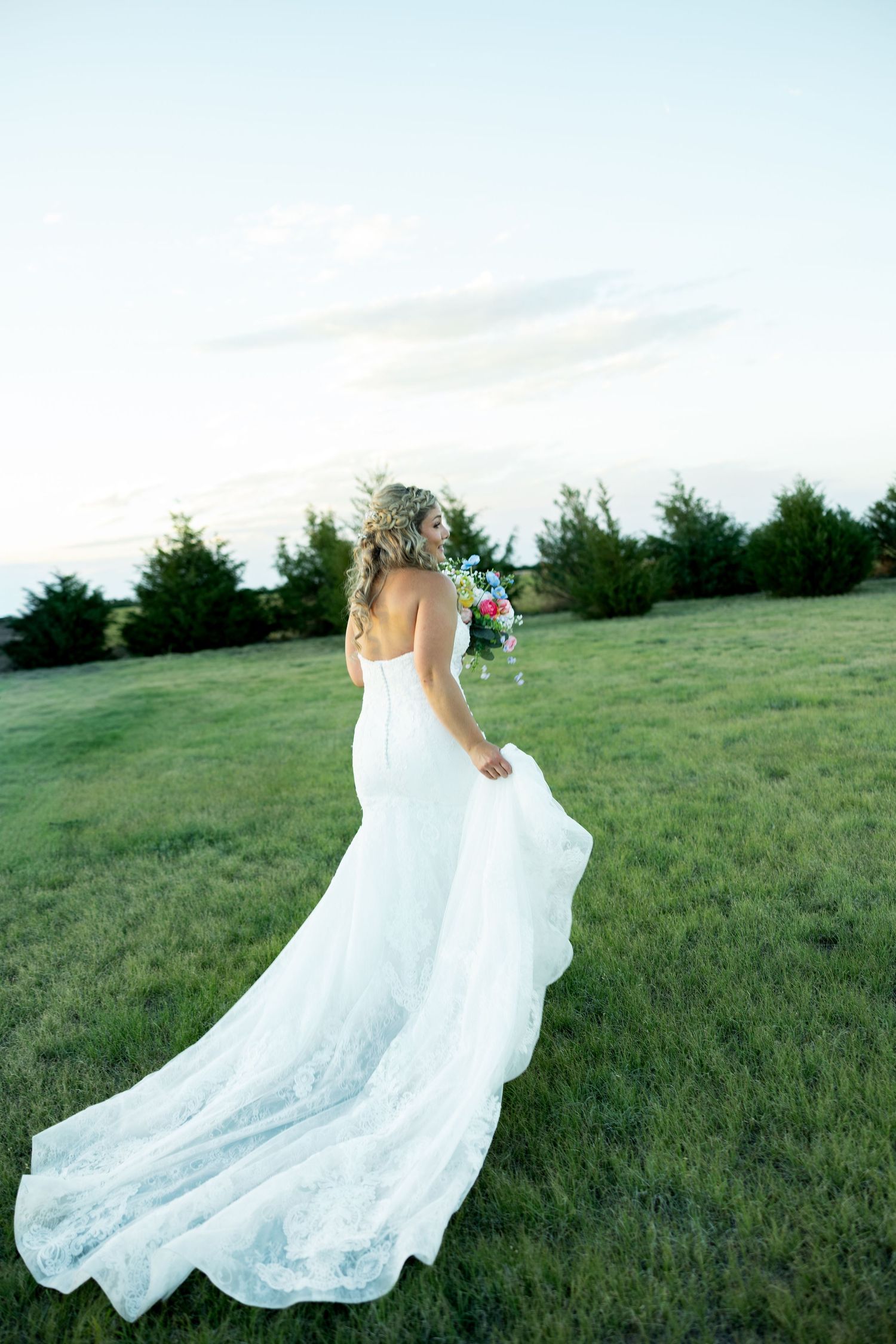 Bride in a flowing white lace gown holds bouquet while standing in a green field at dusk.