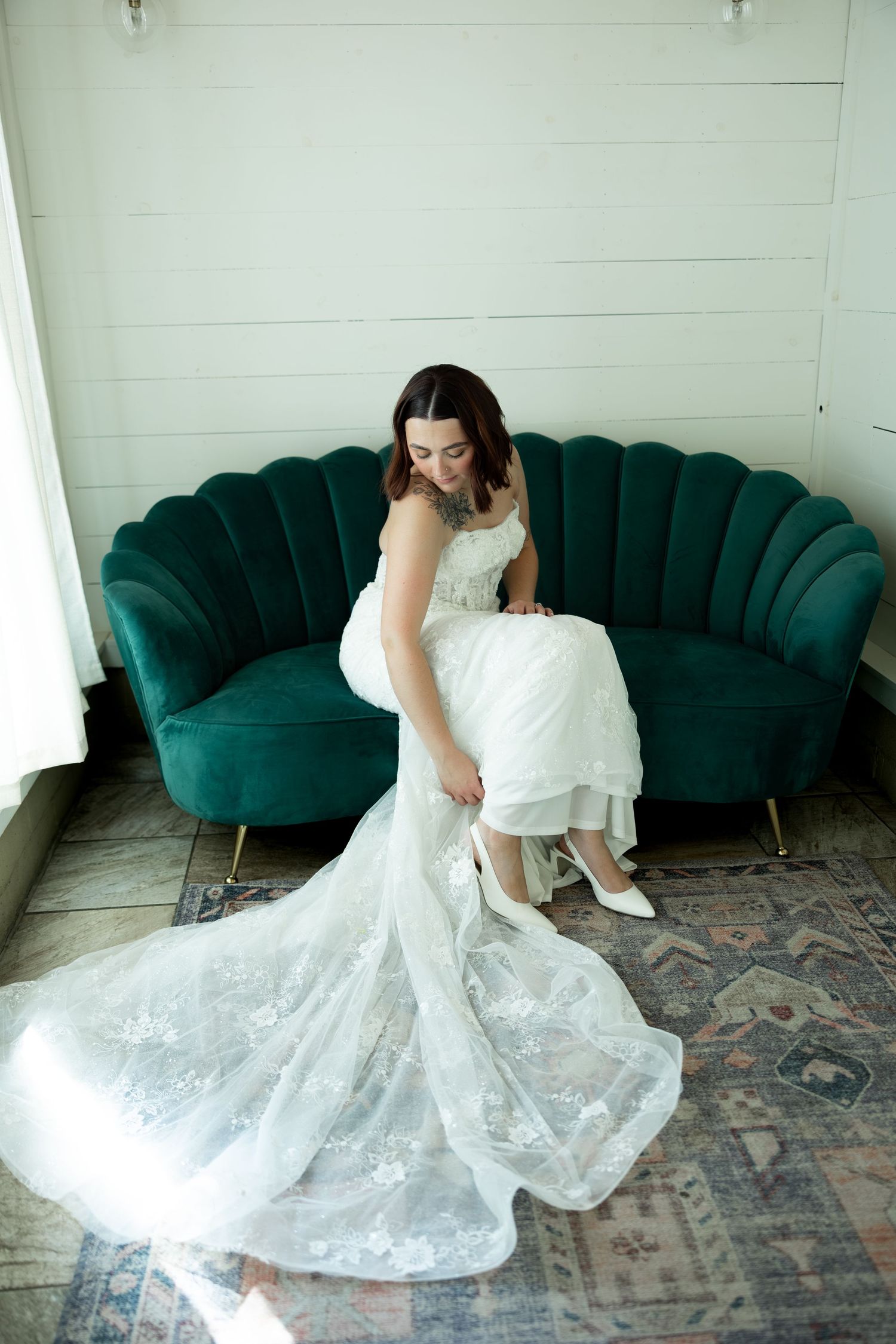Bride in lace wedding gown adjusting white heels while seated on emerald green velvet sofa.