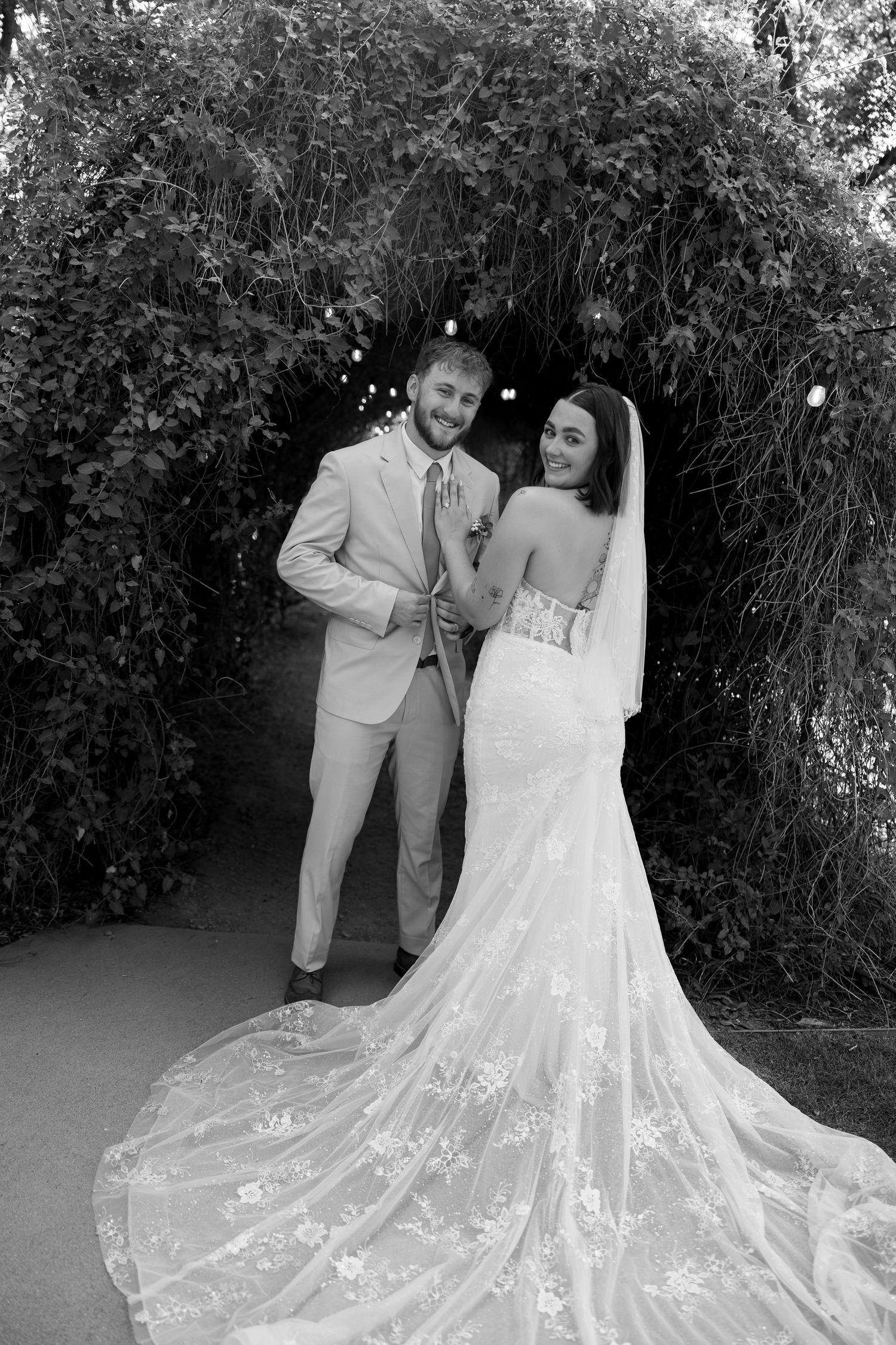 Black and white photo of smiling groom in light suit with bride in lace gown under leafy arch.
