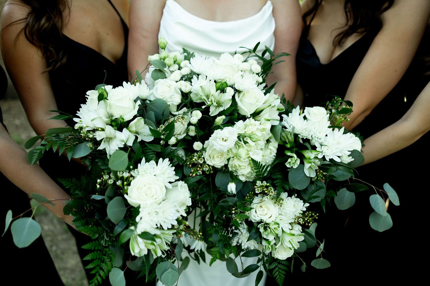 Bride holding lush white floral bouquet with greenery flanked by bridesmaids in black dresses.