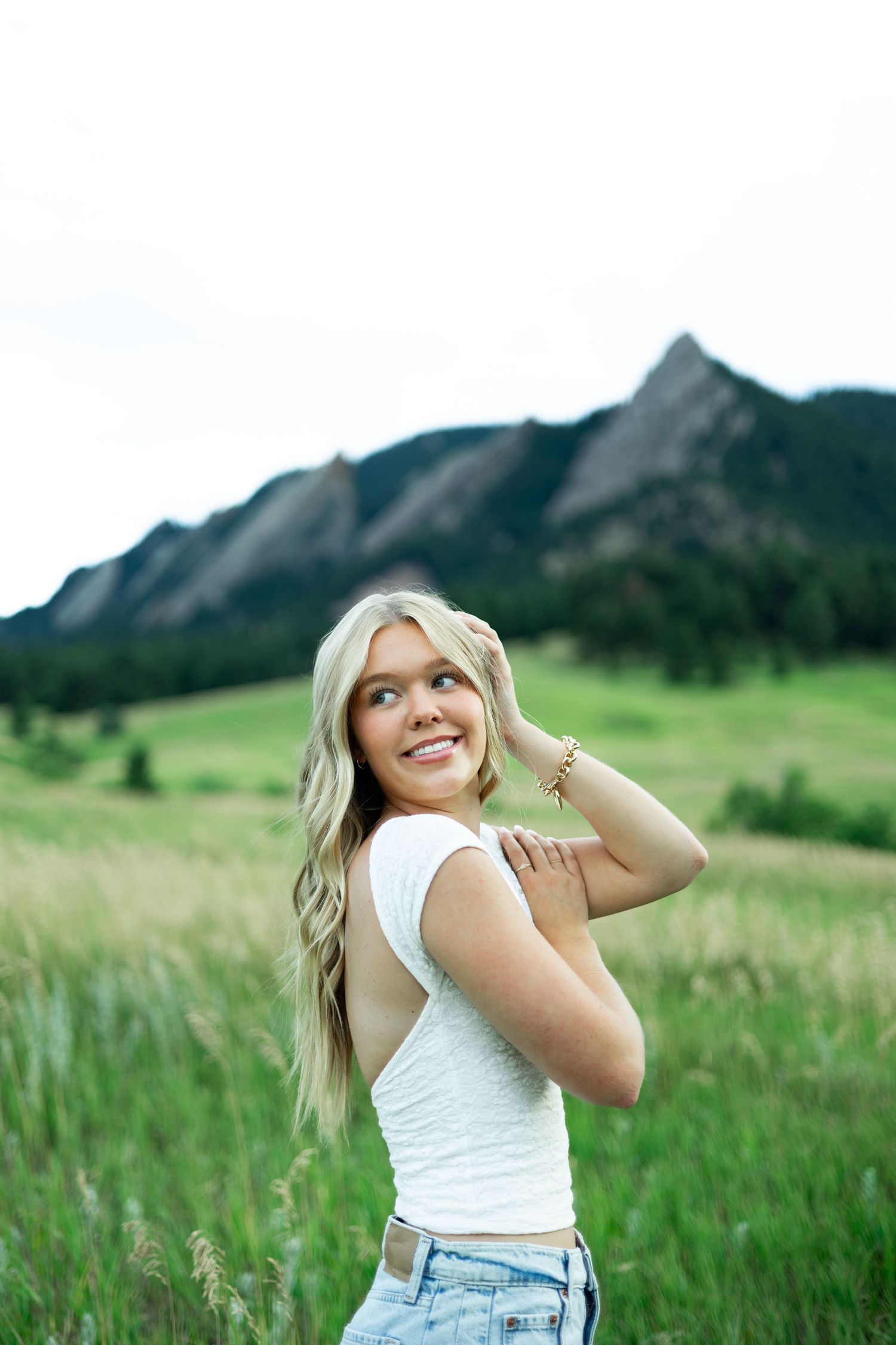 A person in a white top poses playfully in a grassy meadow with dramatic mountain peaks in the background.