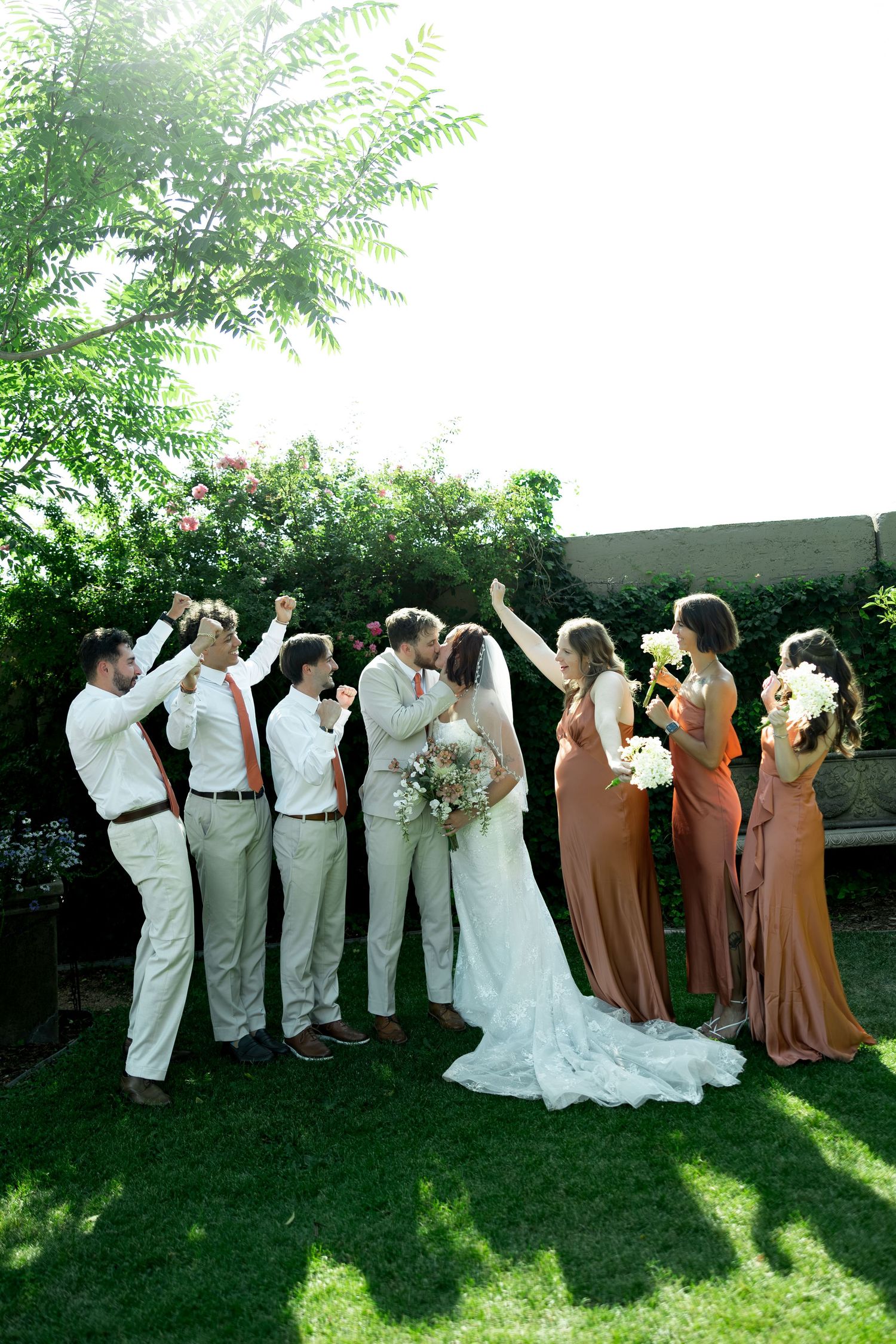 Wedding party celebrates as bride and groom kiss outdoors, bridesmaids in rust dresses and groomsmen in tan suits.
