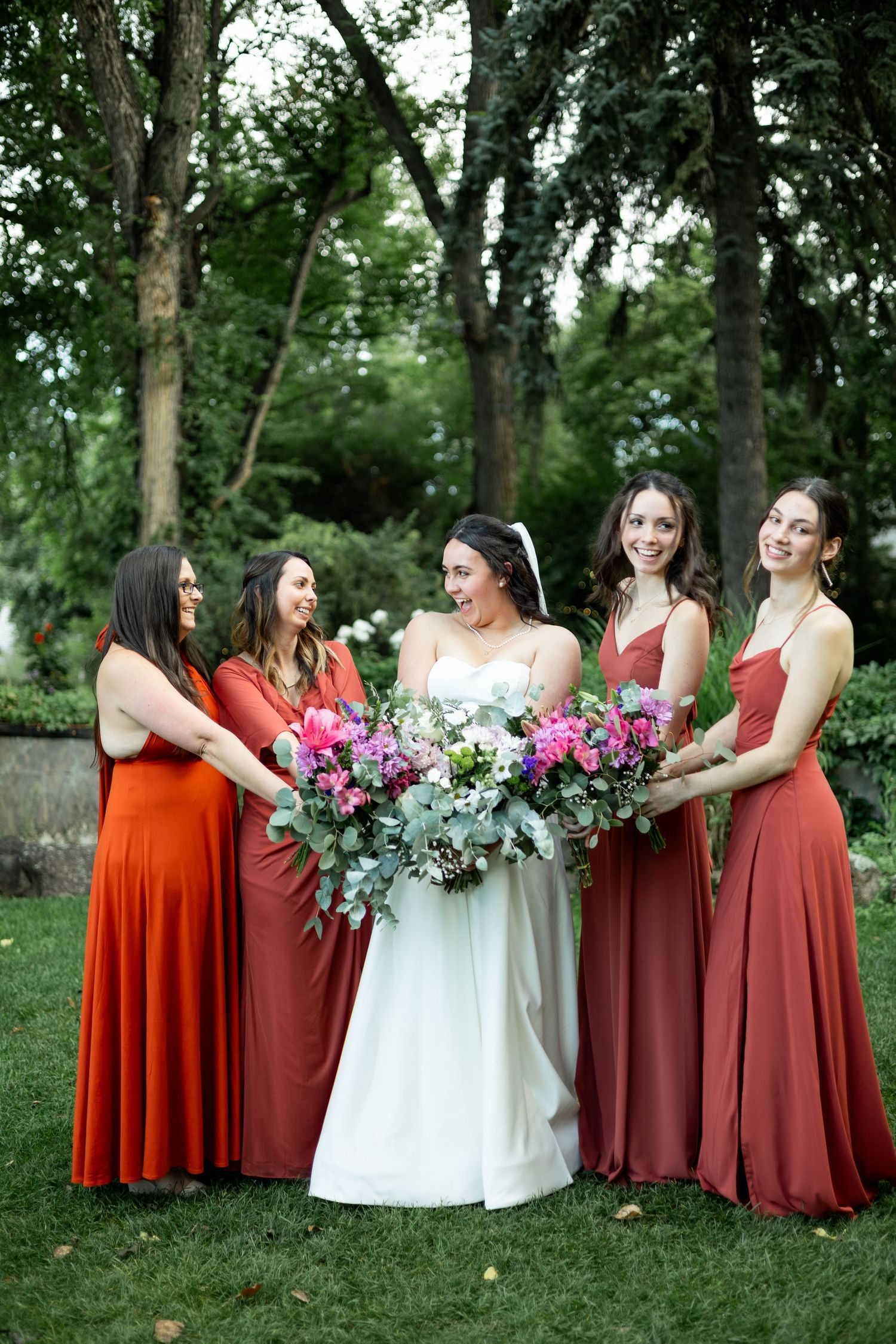 Bridal party in burgundy dresses gather around a bride in white holding colorful bouquets.