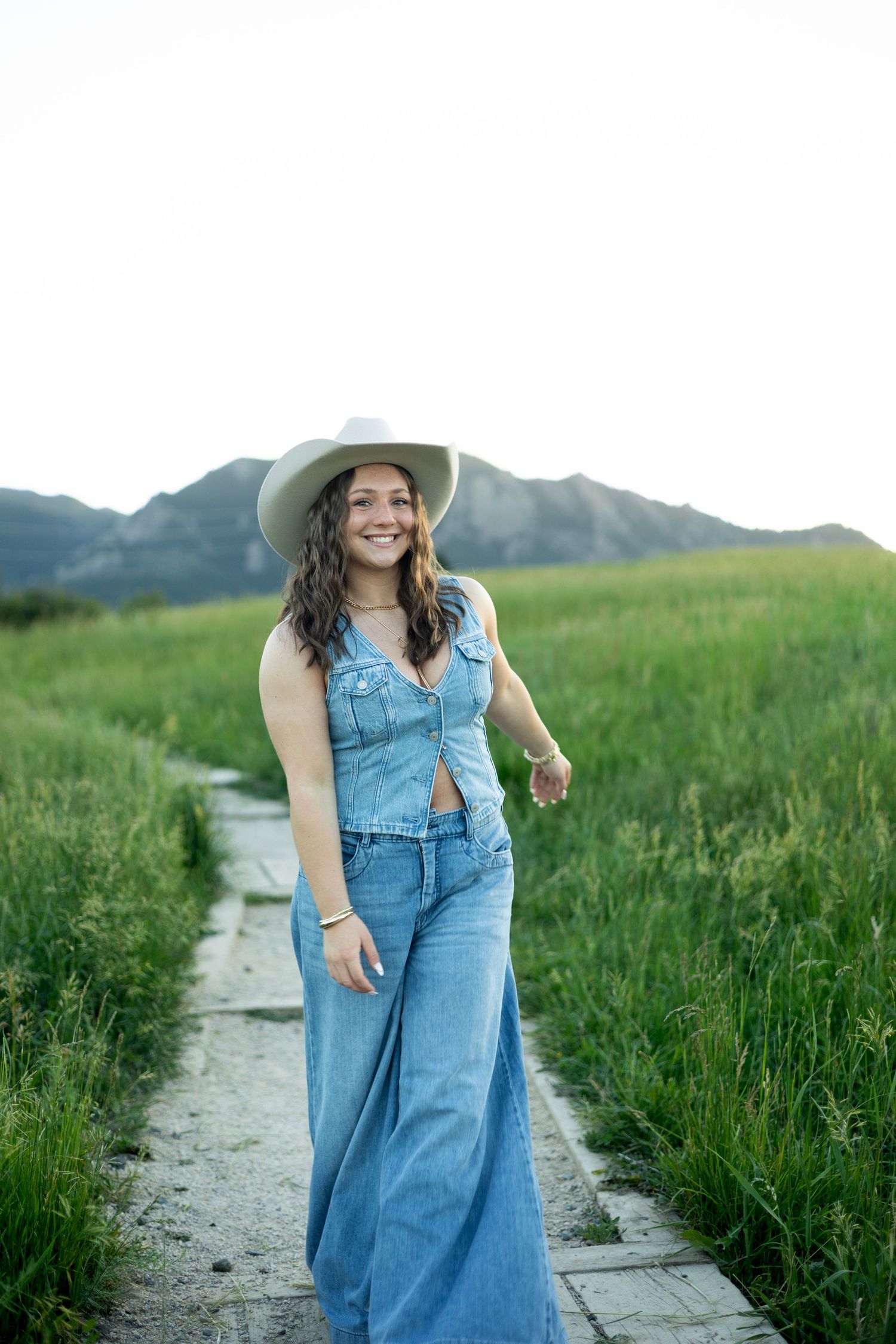 Someone wearing denim overalls and a wide-brim hat walks along a mountain trail surrounded by green grass.