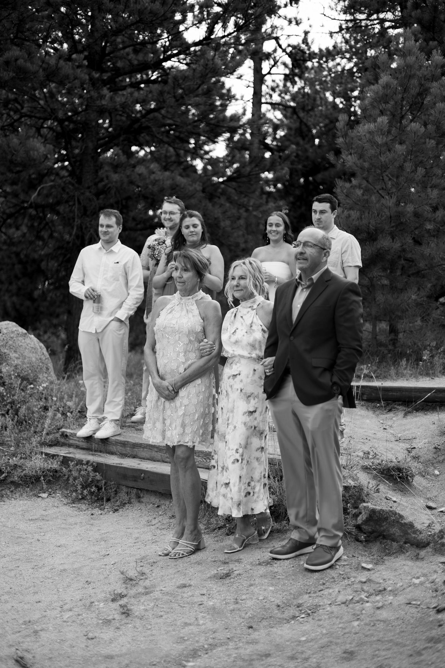 A group poses together on a sandy slope against a backdrop of trees in casual and summer attire.