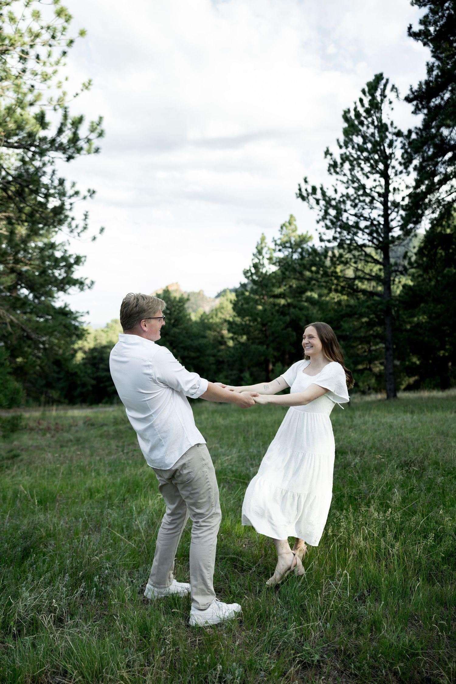 Couple in white outfits dancing joyfully in a green meadow surrounded by pine trees on a cloudy day.