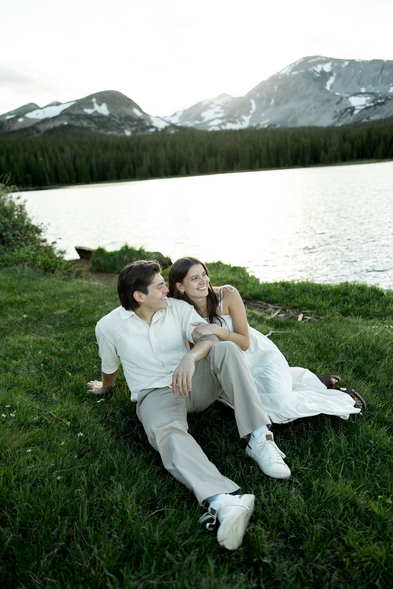 A happy couple sits together on green grass by a serene mountain lake, smiling and relaxing in nature.