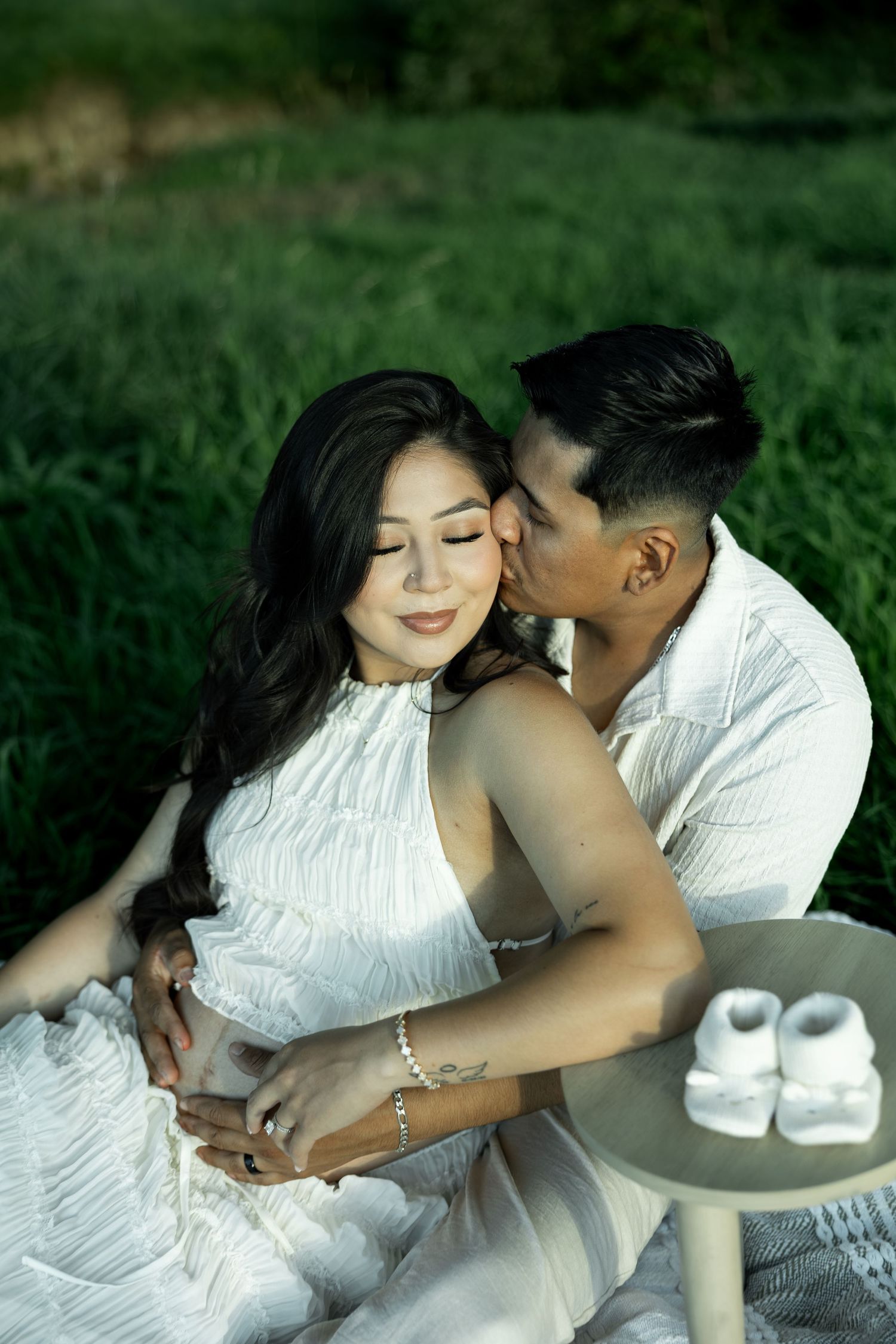 A couple in white outfits sit in a green field, man kissing woman's cheek, tiny baby shoes nearby.