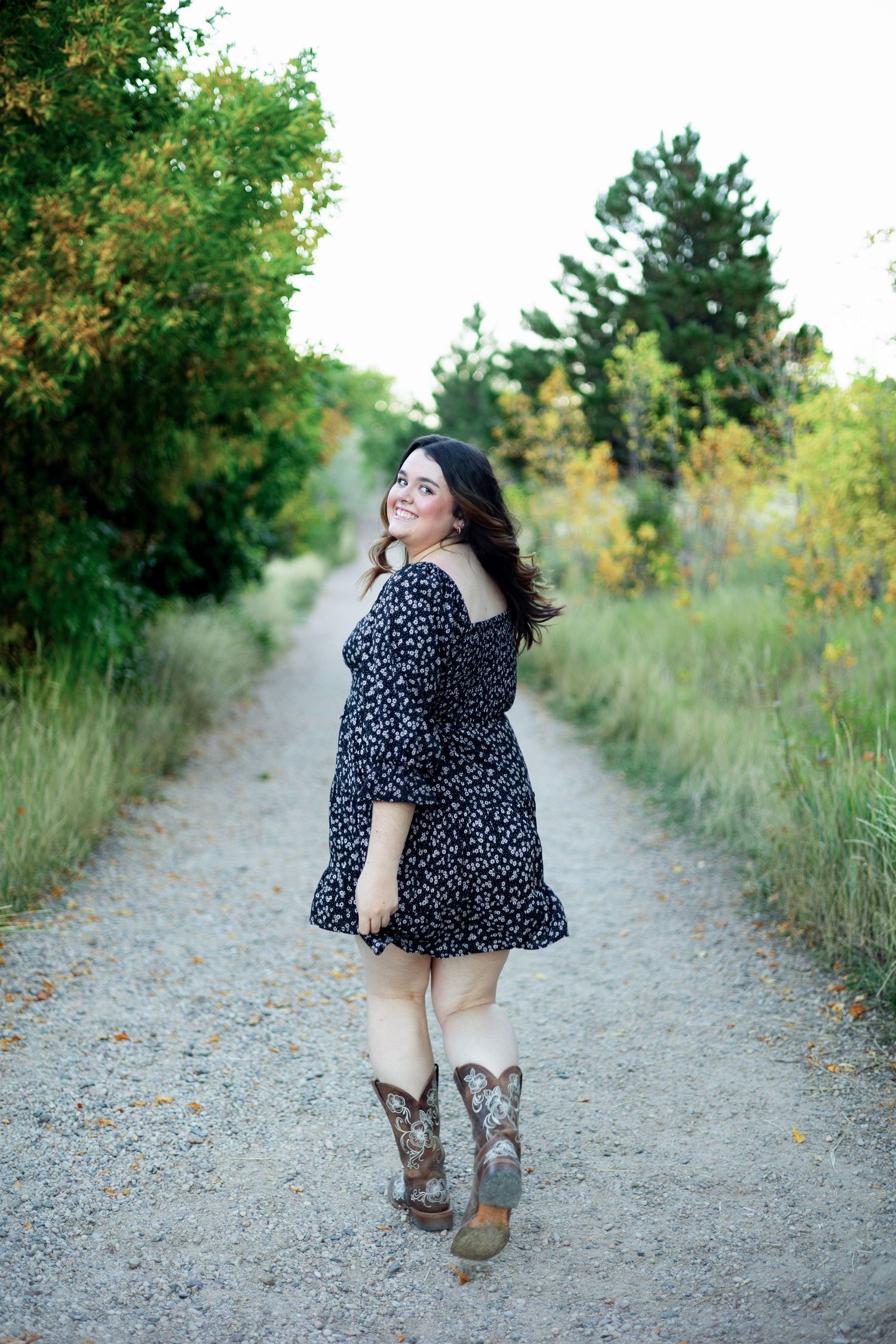 Young woman in floral dress and cowboy boots smiling on a scenic gravel path surrounded by lush greenery.