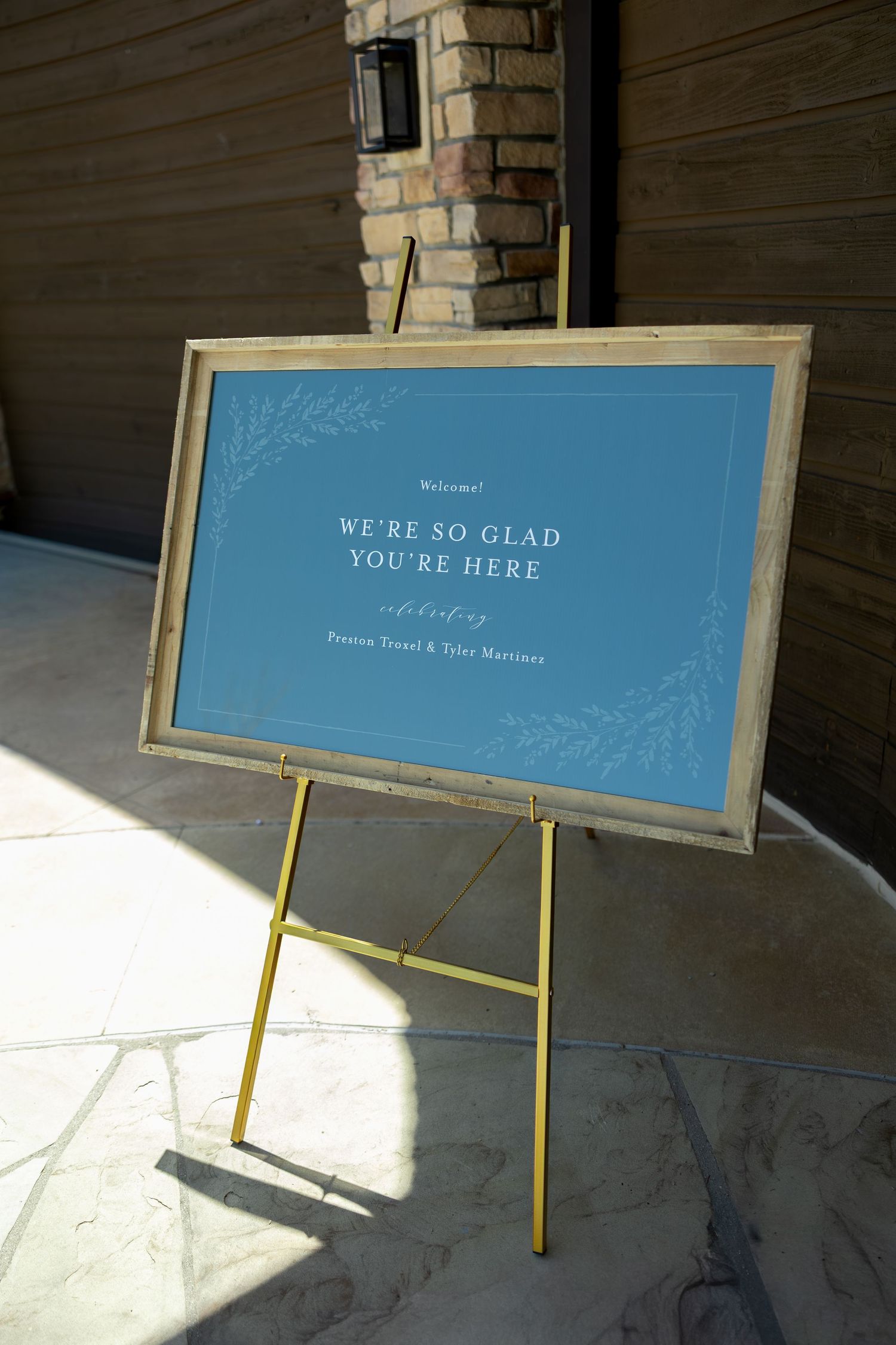 Blue wedding welcome sign on gold easel reads We're So Glad You're Here celebrating Preston Tranel and Tyler Martinez.