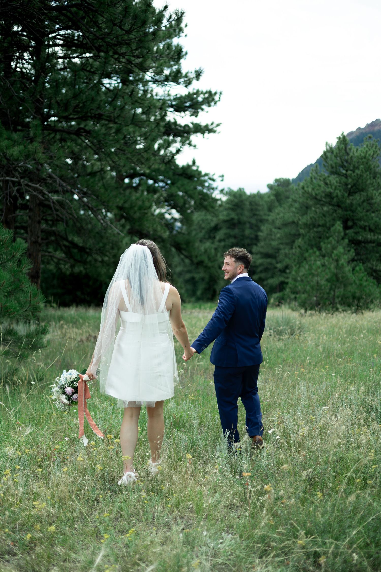 A couple in wedding attire playfully picks wildflowers together in a scenic meadow surrounded by pine trees.