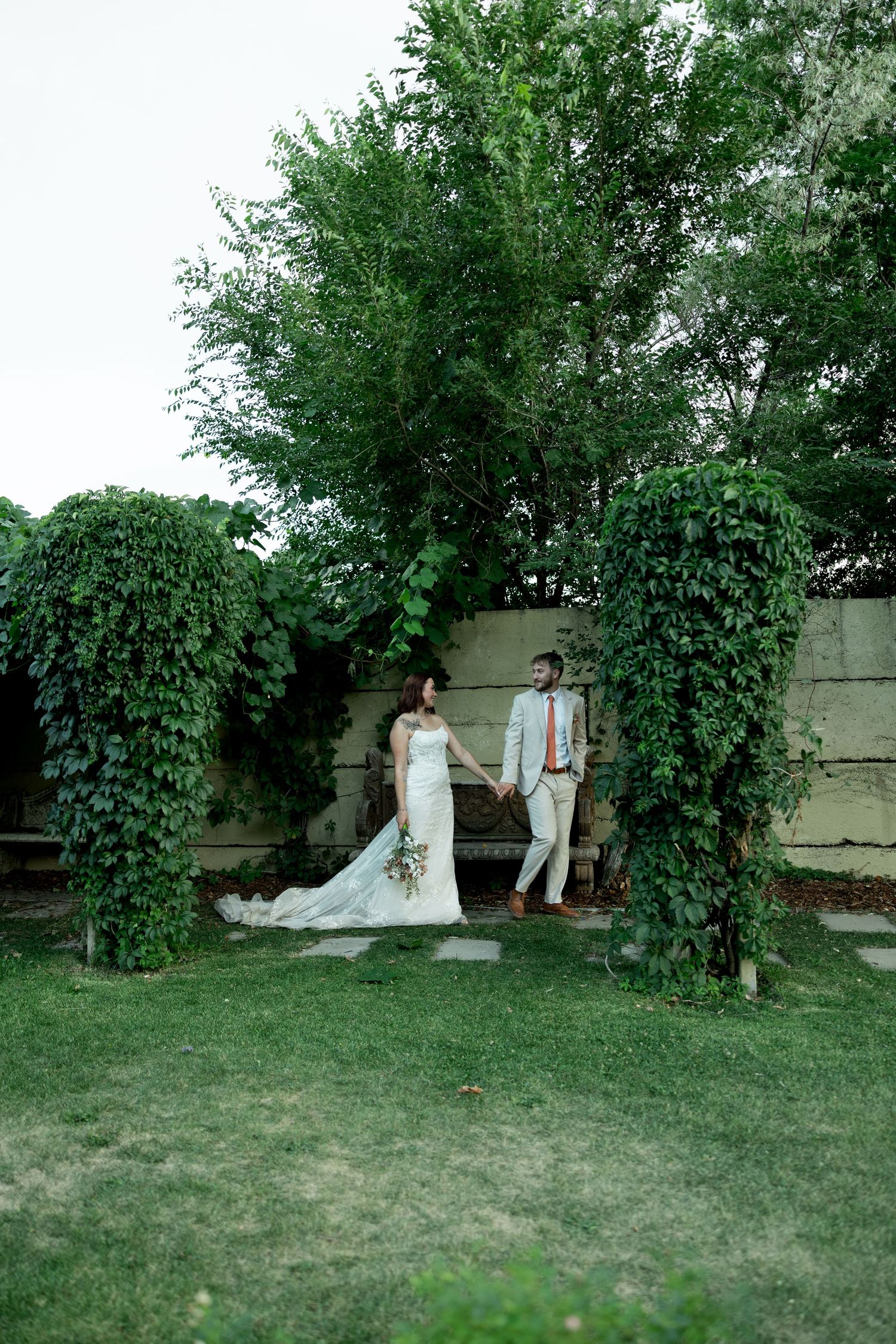 Bride and groom hold hands in lush ivy-covered garden archway during outdoor wedding portrait.