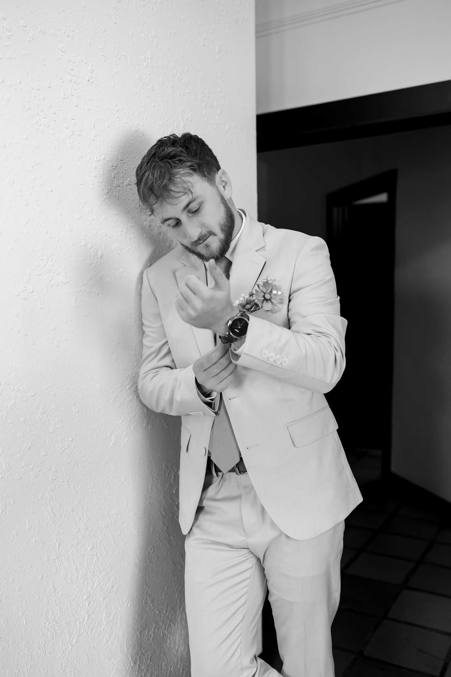 A dapper groom in a light suit adjusts his watch while leaning against a white wall in a black and white photo.