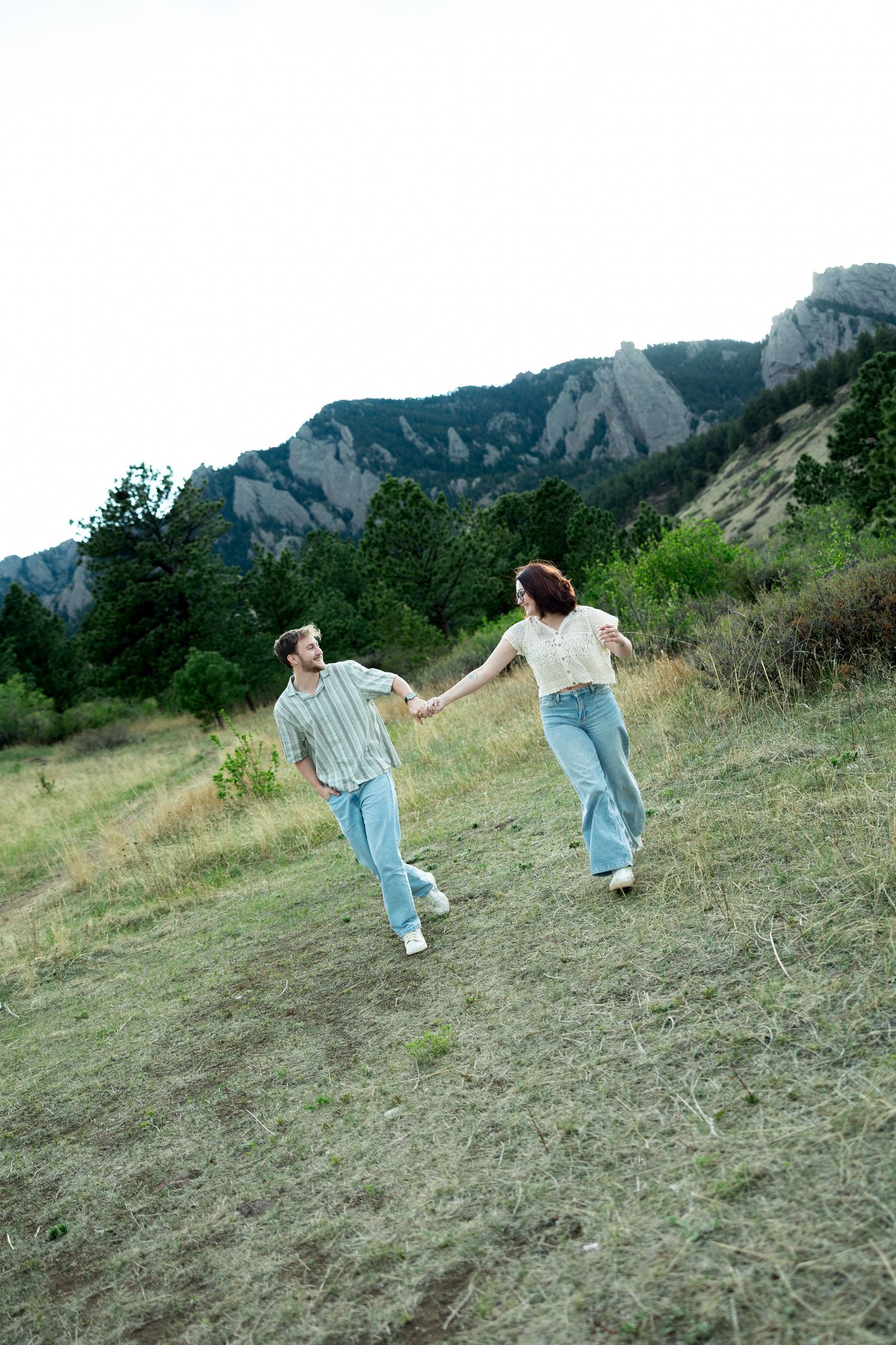 A couple holds hands while running through a grassy mountain meadow, both wearing jeans and light tops.