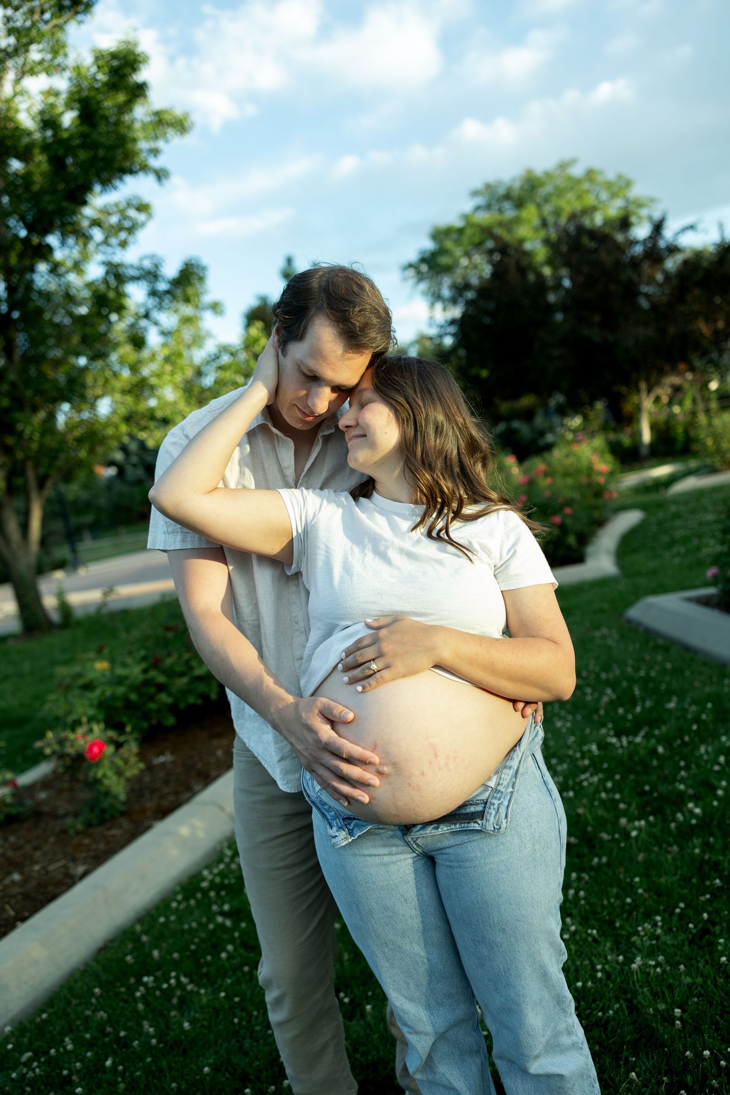 Happy couple in park, pregnant woman in white top and jeans embraced by partner while cradling baby bump.
