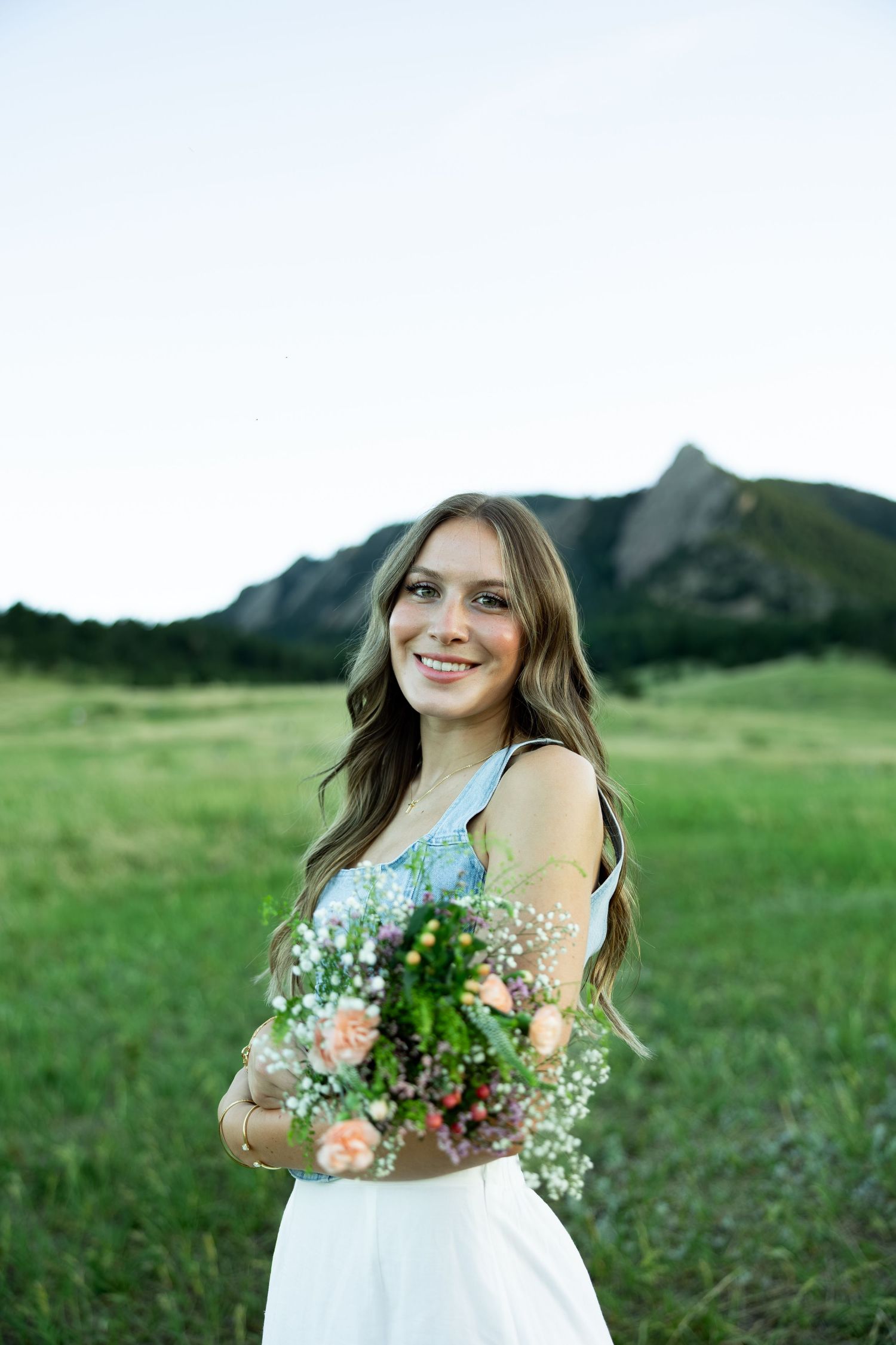 Smiling young woman holding a wildflower bouquet in a green meadow with mountain peaks in the background.