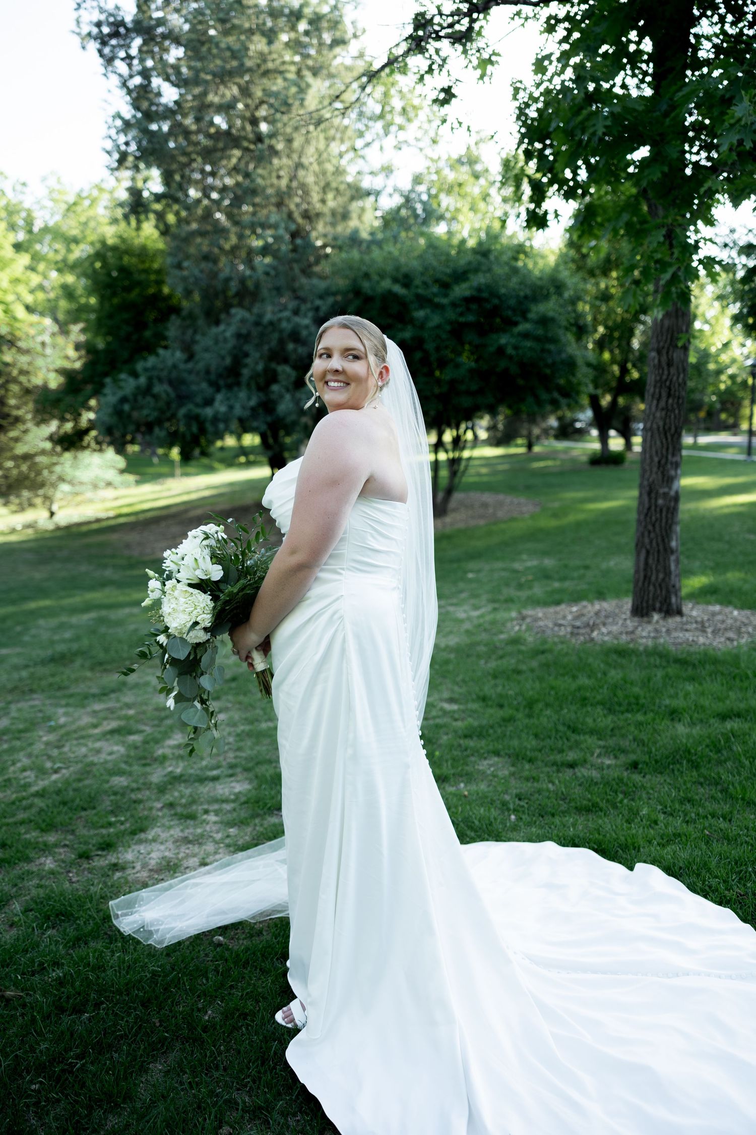 Smiling bride in white gown with train and veil holds bouquet of white flowers while posing in a lush green park.