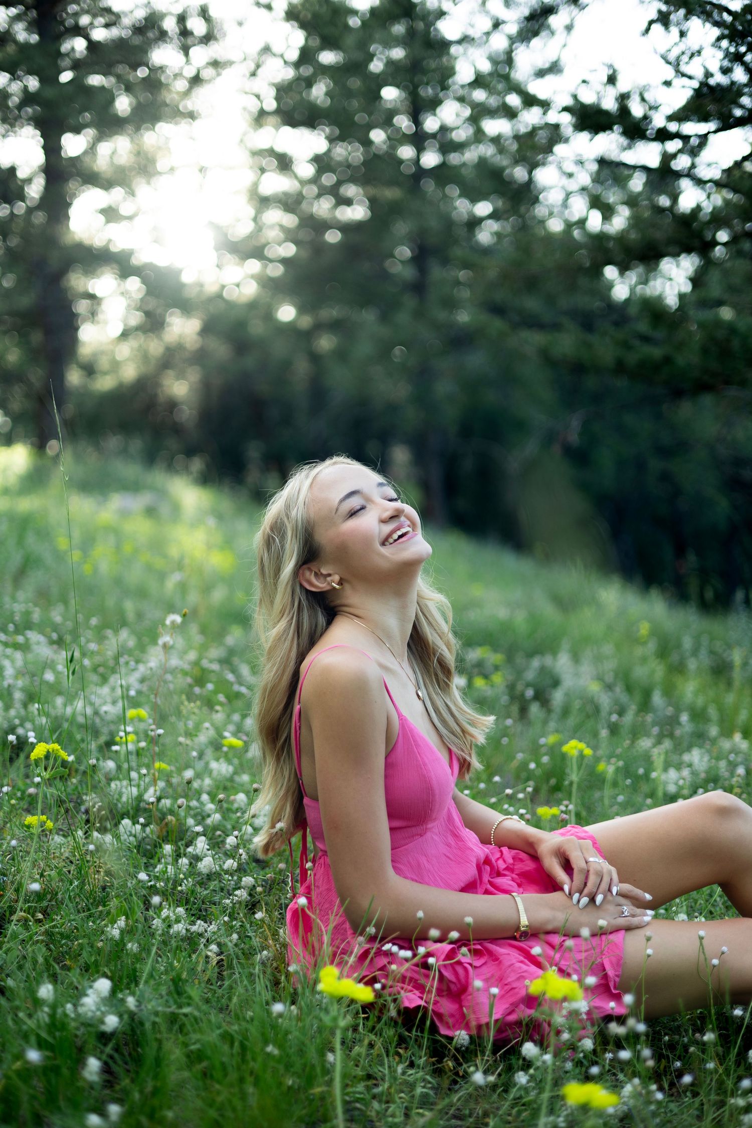 Young blonde woman in pink dress sitting joyfully in wildflower meadow surrounded by lush green trees.