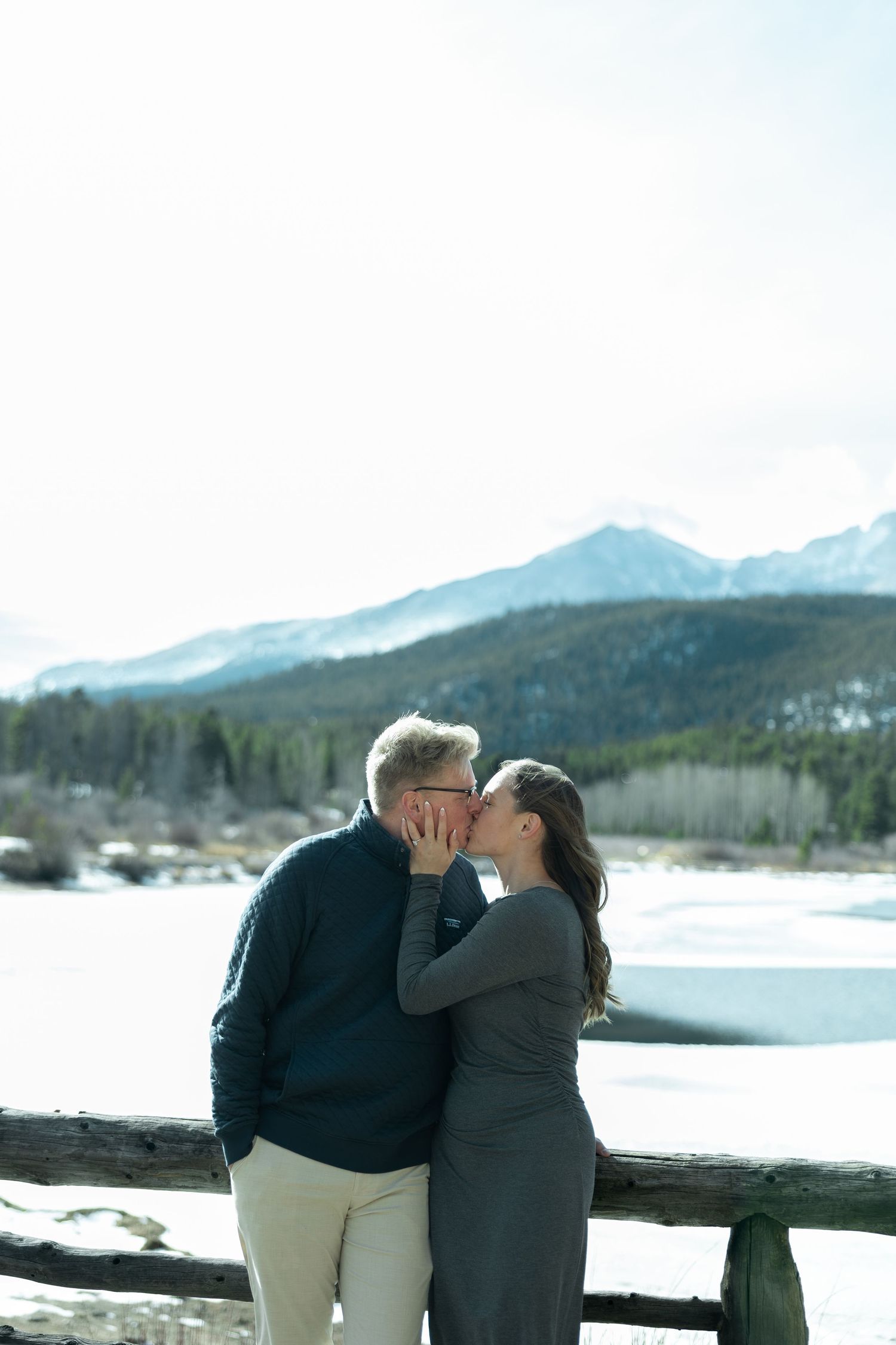 Couple sharing a romantic kiss by a rustic fence with snowy mountains and frozen lake backdrop.