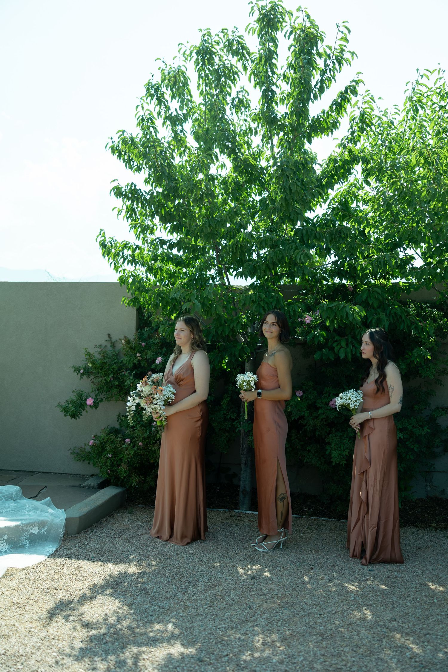 Three bridesmaids in rust-colored satin gowns holding white bouquets stand outdoors near a lush green tree.