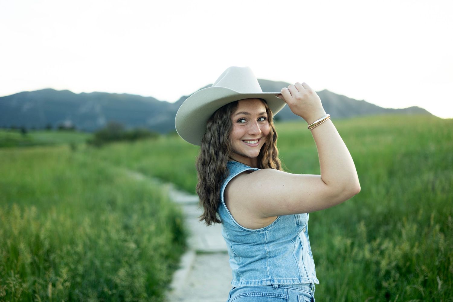A person wearing a denim dress and hat walks along a winding path through lush green summer fields.