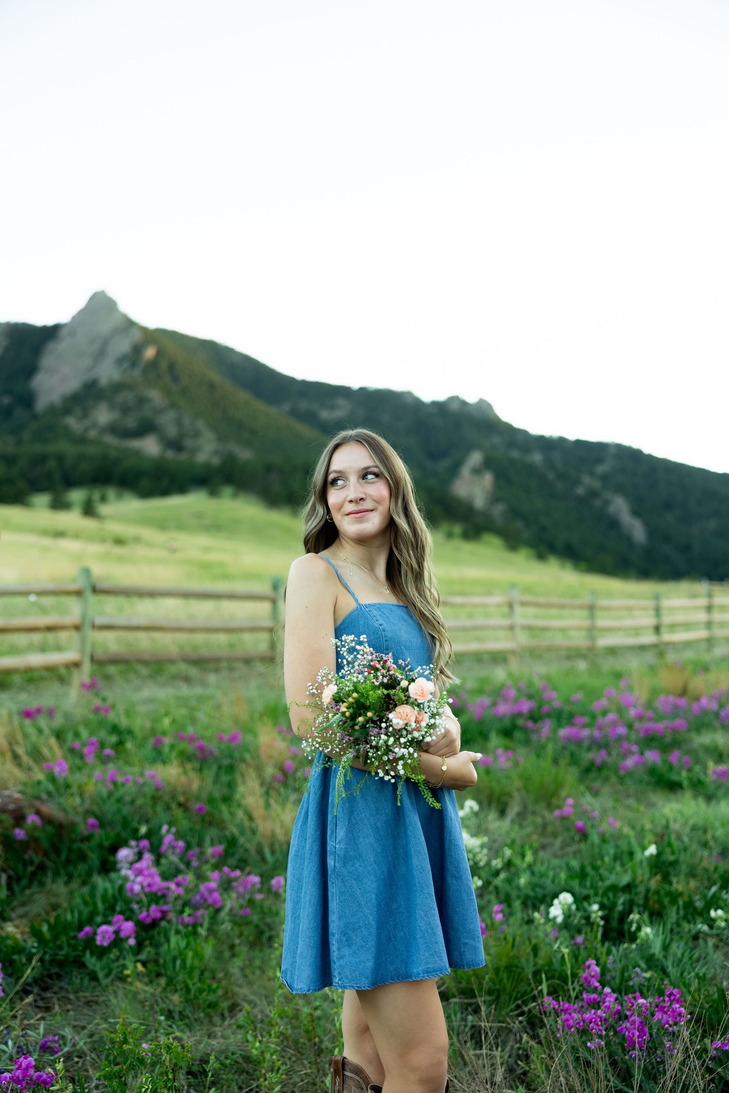Young woman in blue dress holding wildflower bouquet standing in field of purple flowers near mountains.