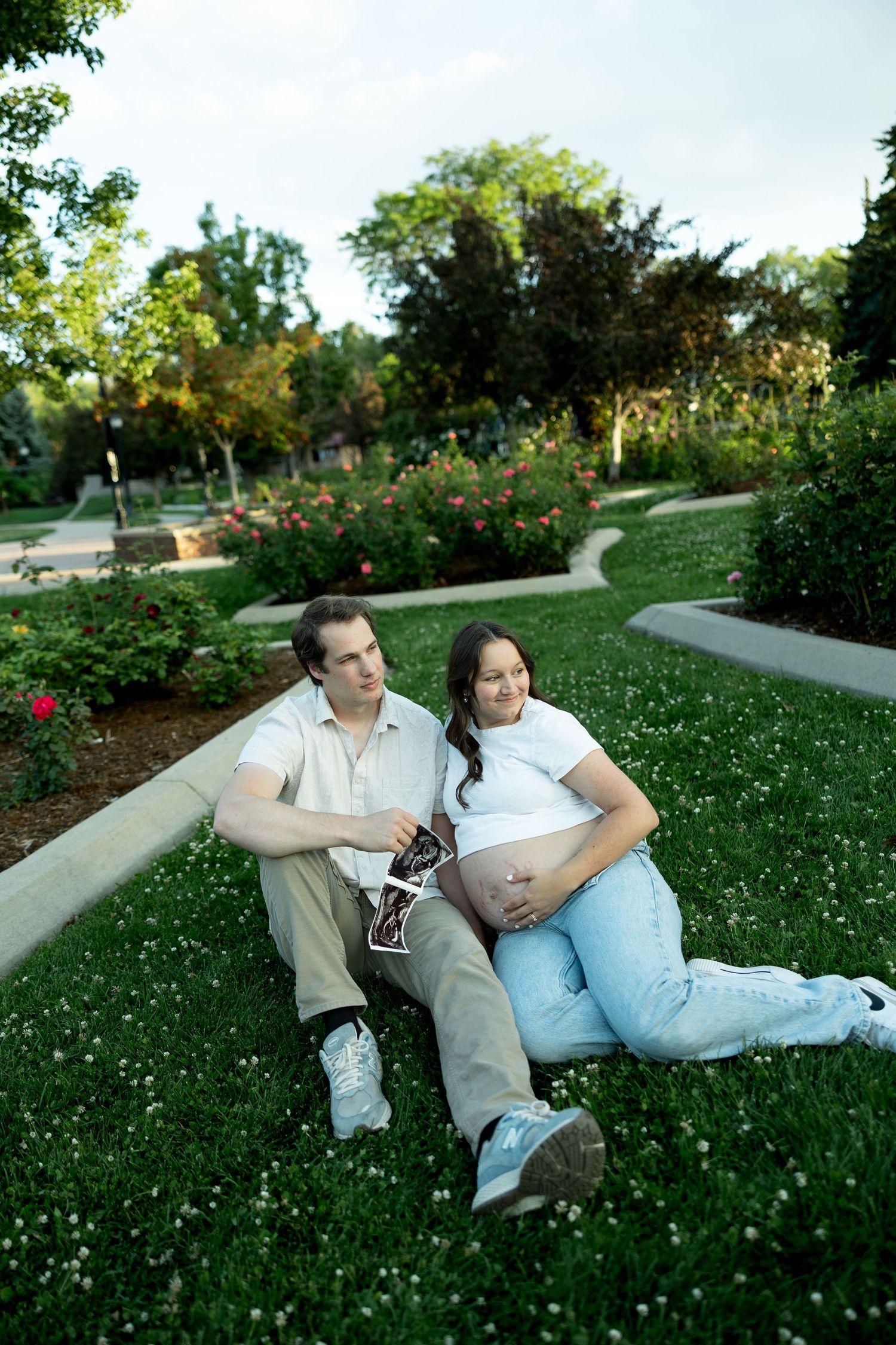 Pregnant couple sitting on grass in a park, holding ultrasound photos, sharing a tender moment outdoors.