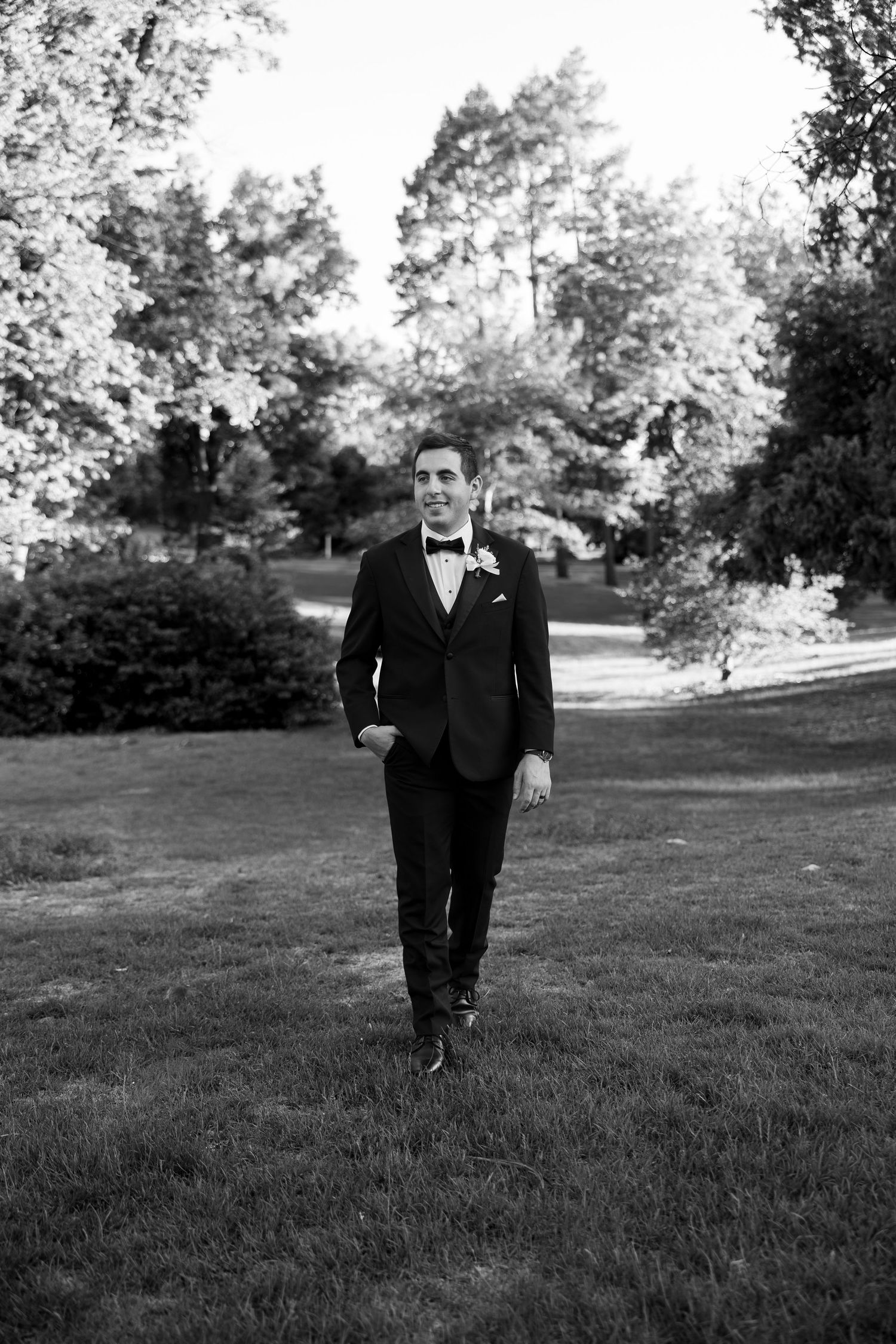 Groom in black tuxedo with bow tie walks confidently across green lawn in black and white photo.
