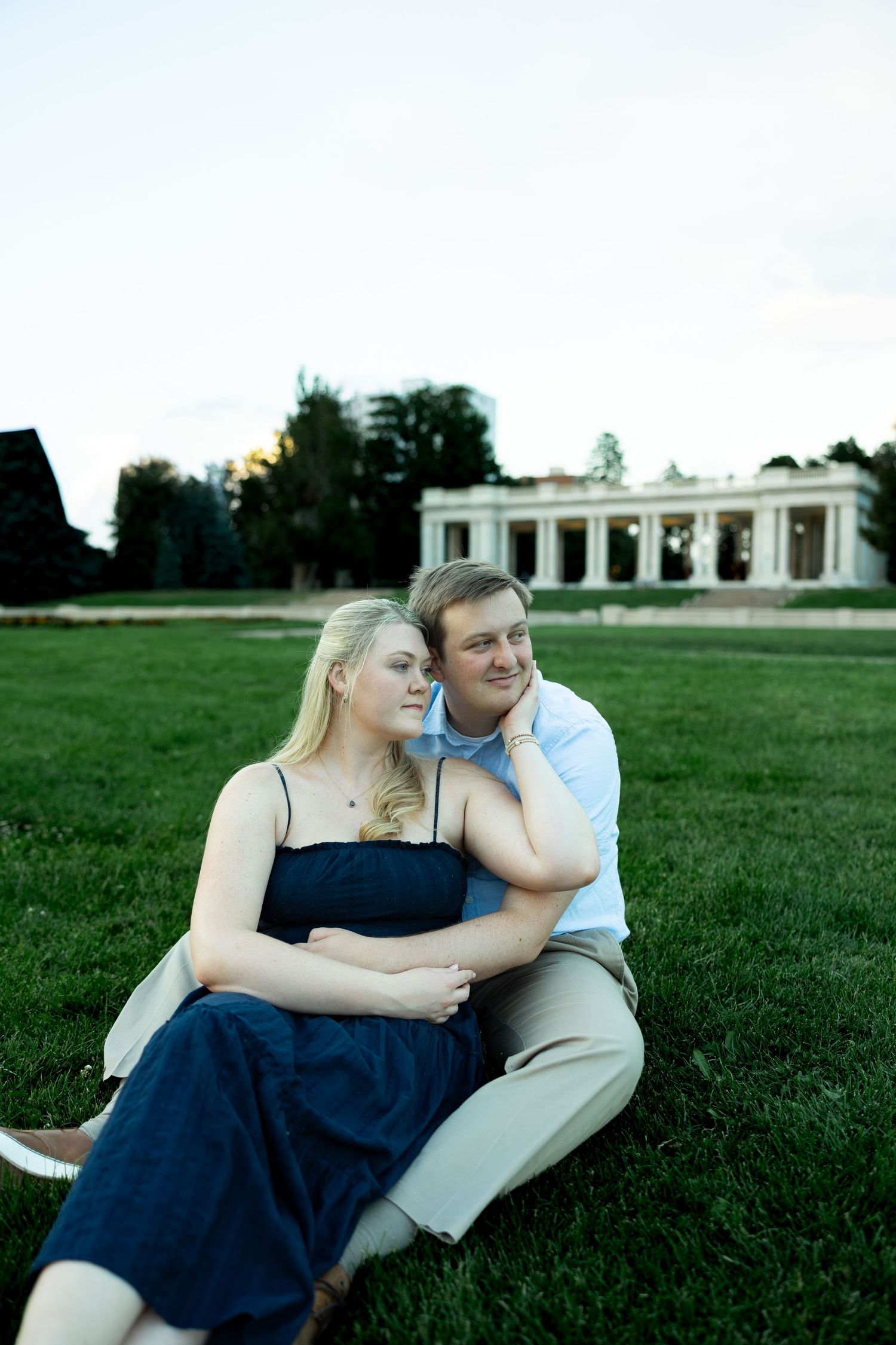 Couple sitting on green grass in front of a classical building, woman in navy dress leaning against man in light blue shirt.